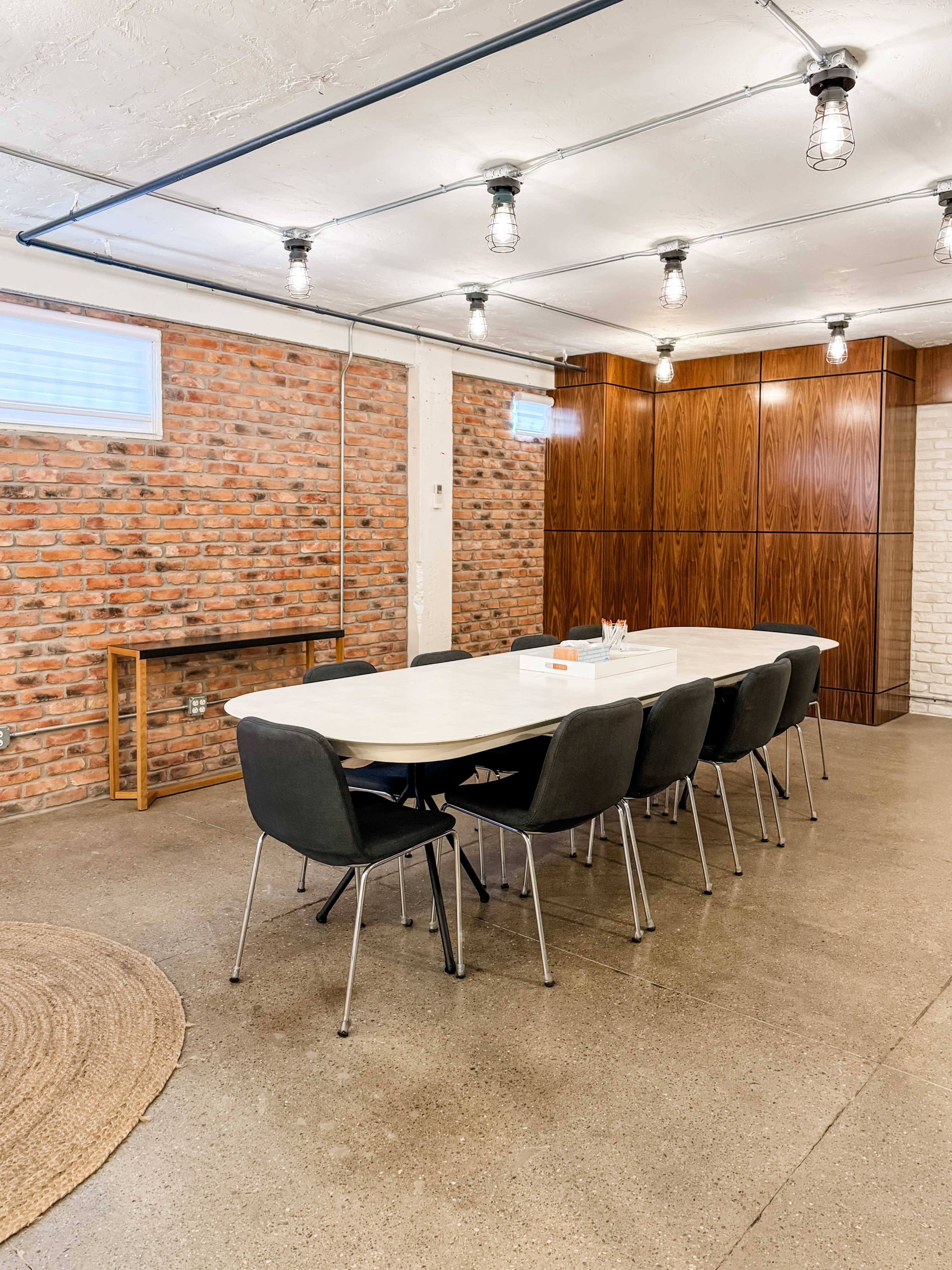 A long white table with black chairs is set up in a room featuring exposed brick walls and overhead pendant lights.