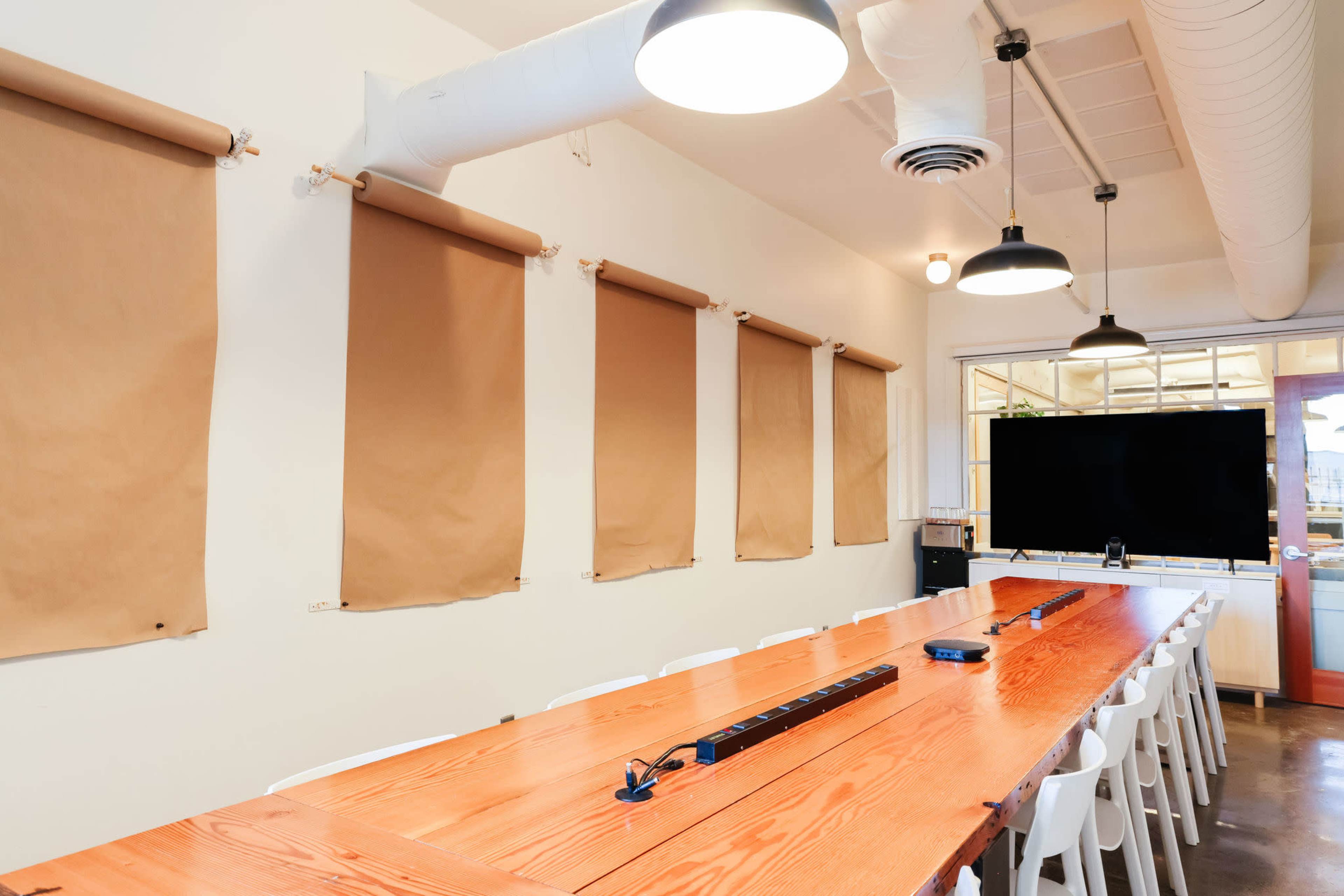 A long wooden table is set in a well-lit meeting room with brown paper roll blinds on the walls and a large screen at one end.