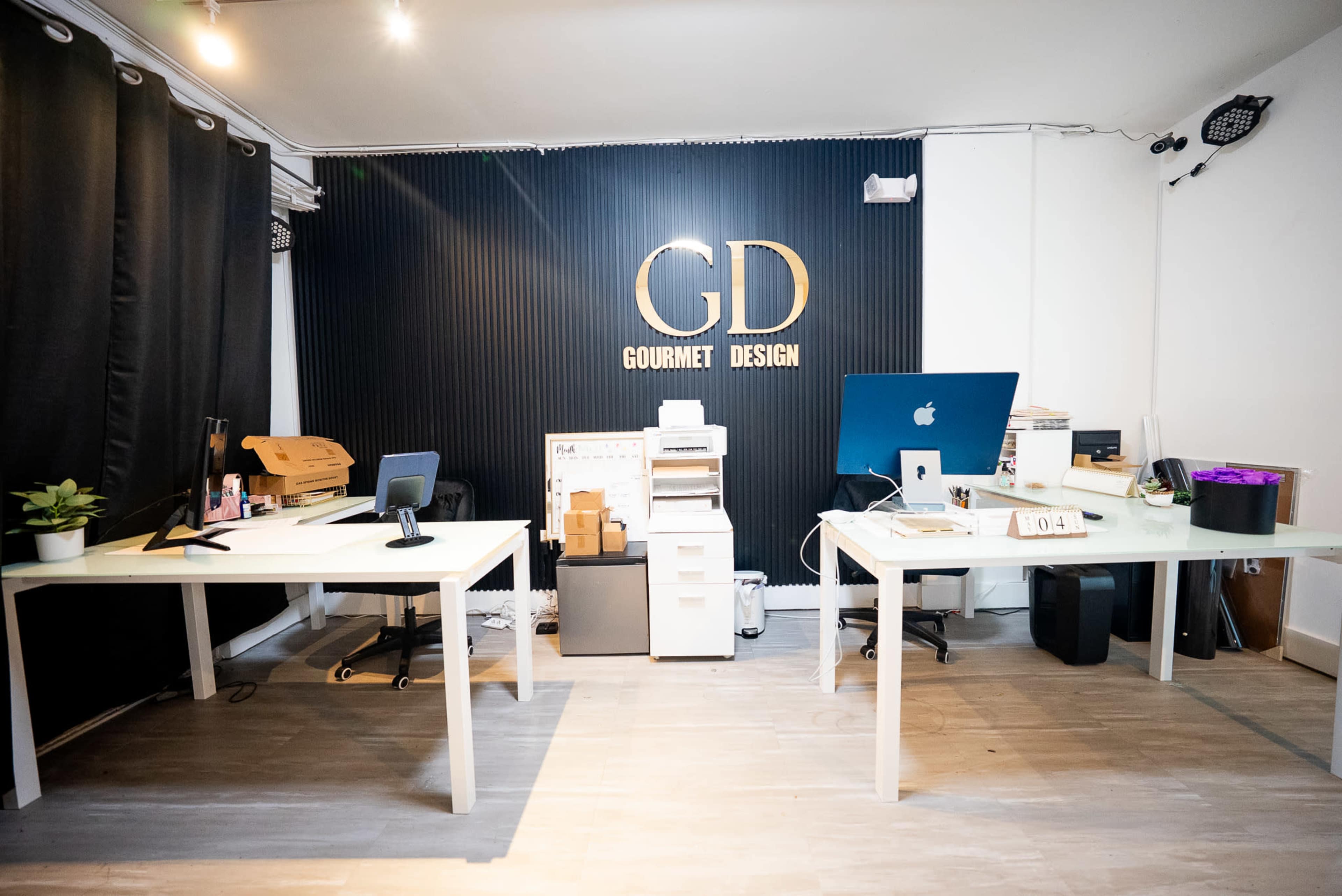 The image shows a modern office space with two desks, a printer, and a wall featuring the name "Gourmet Design" in gold lettering.