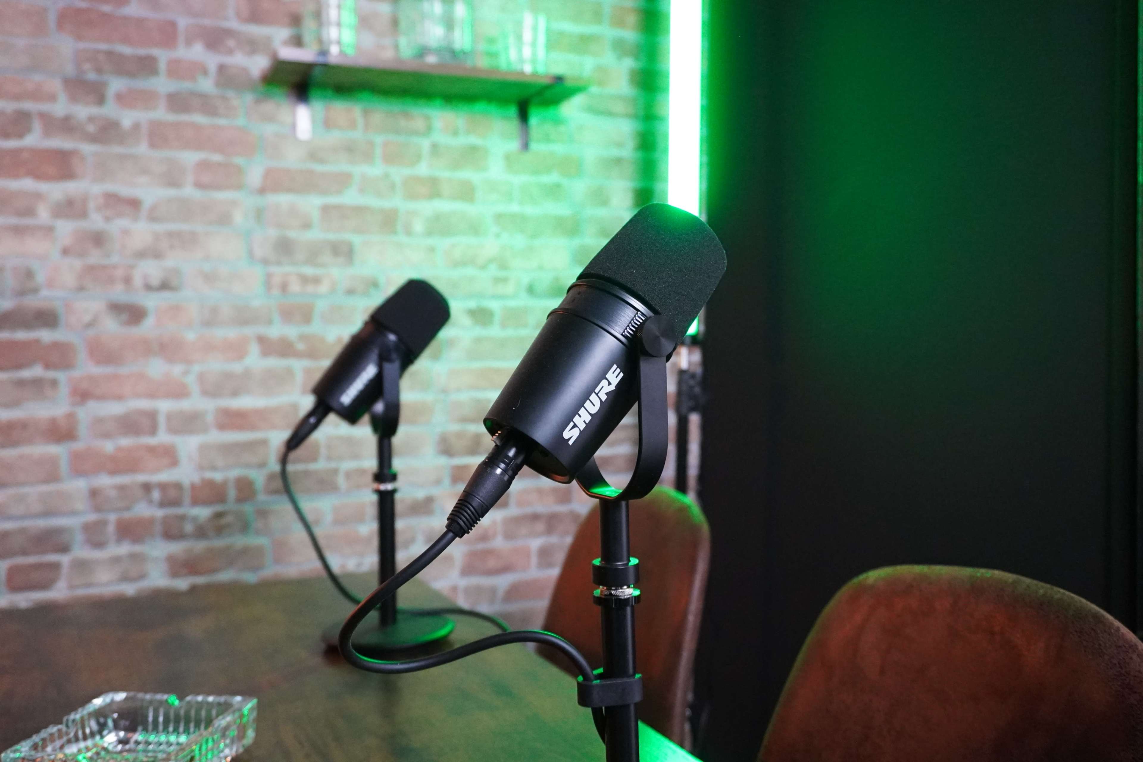 The image shows two microphones mounted on stands on a wooden table in a room with exposed brick walls and green LED lighting.