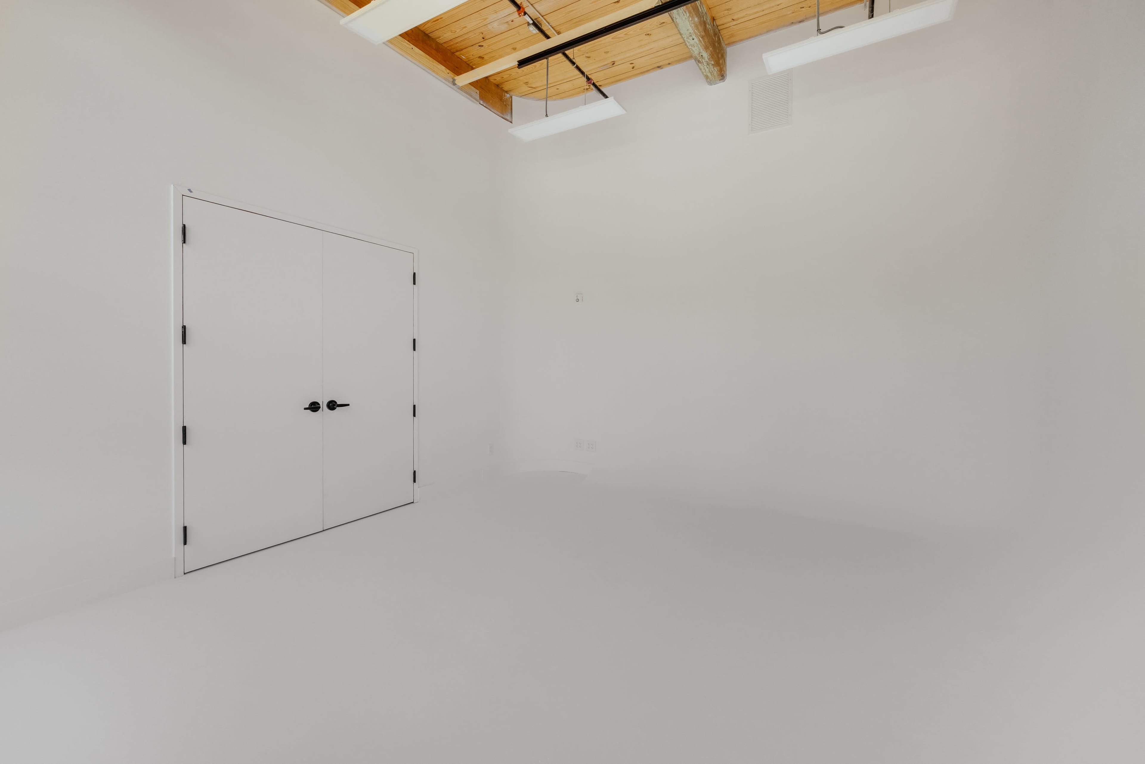 The image shows a minimalist room with white walls and a flat white floor, featuring a closed double door and exposed wooden beams on the ceiling.