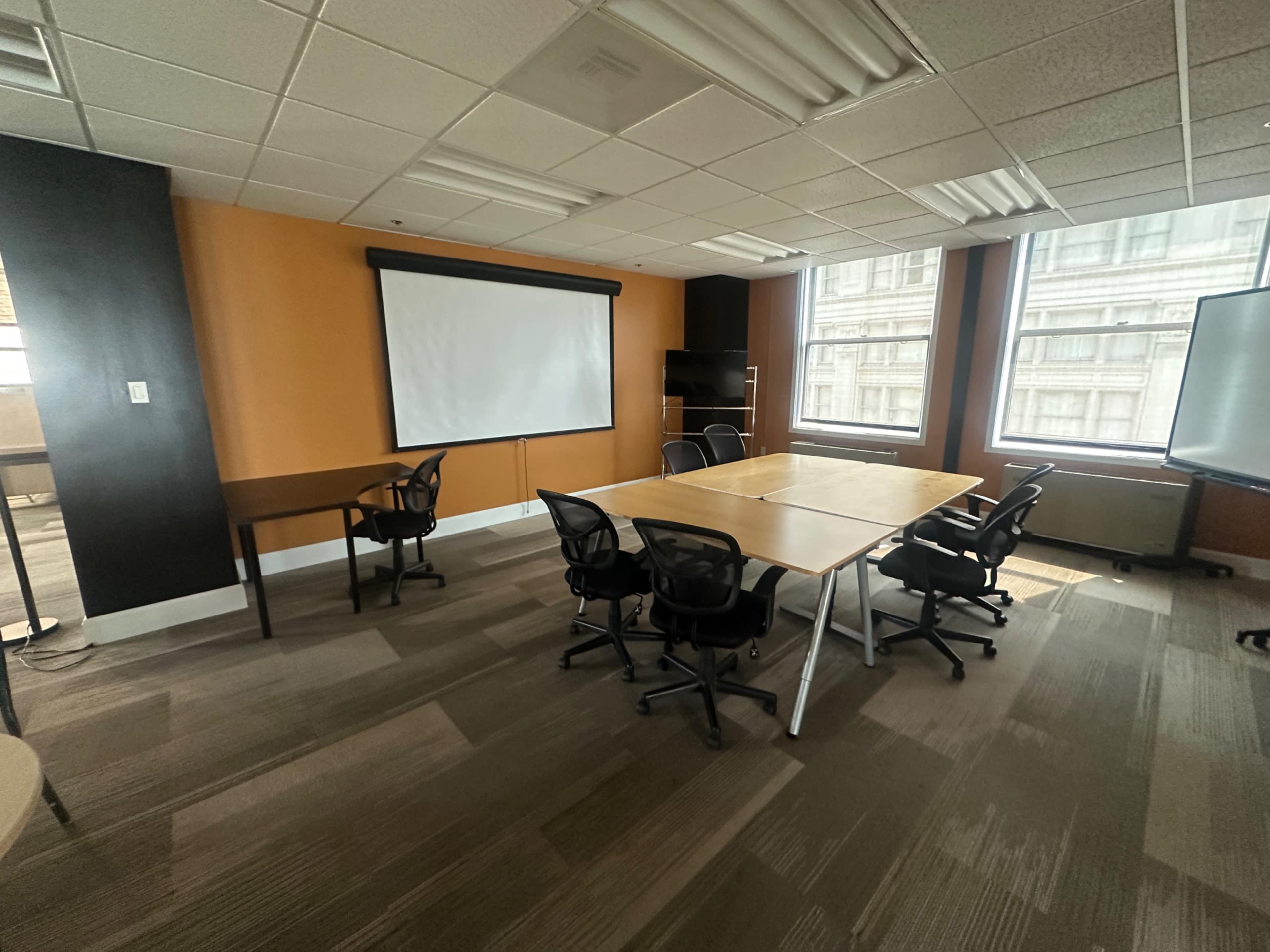 The image shows a conference room featuring a large table surrounded by several chairs, a projector screen at one end, and windows letting in natural light.