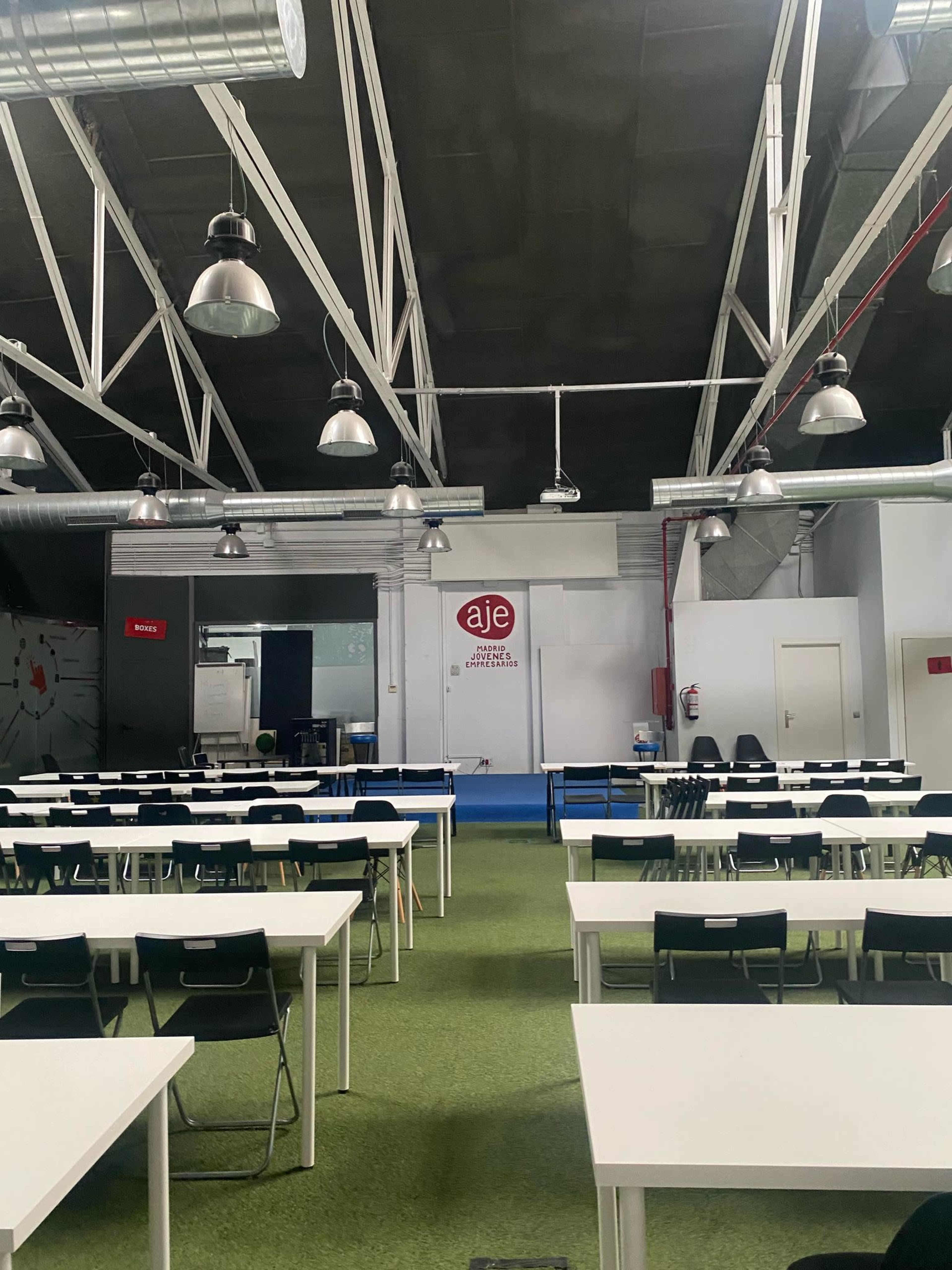 The image shows a spacious room with rows of white tables and chairs, a blue carpet, and industrial-style lighting, featuring a wall with the logo "aje."