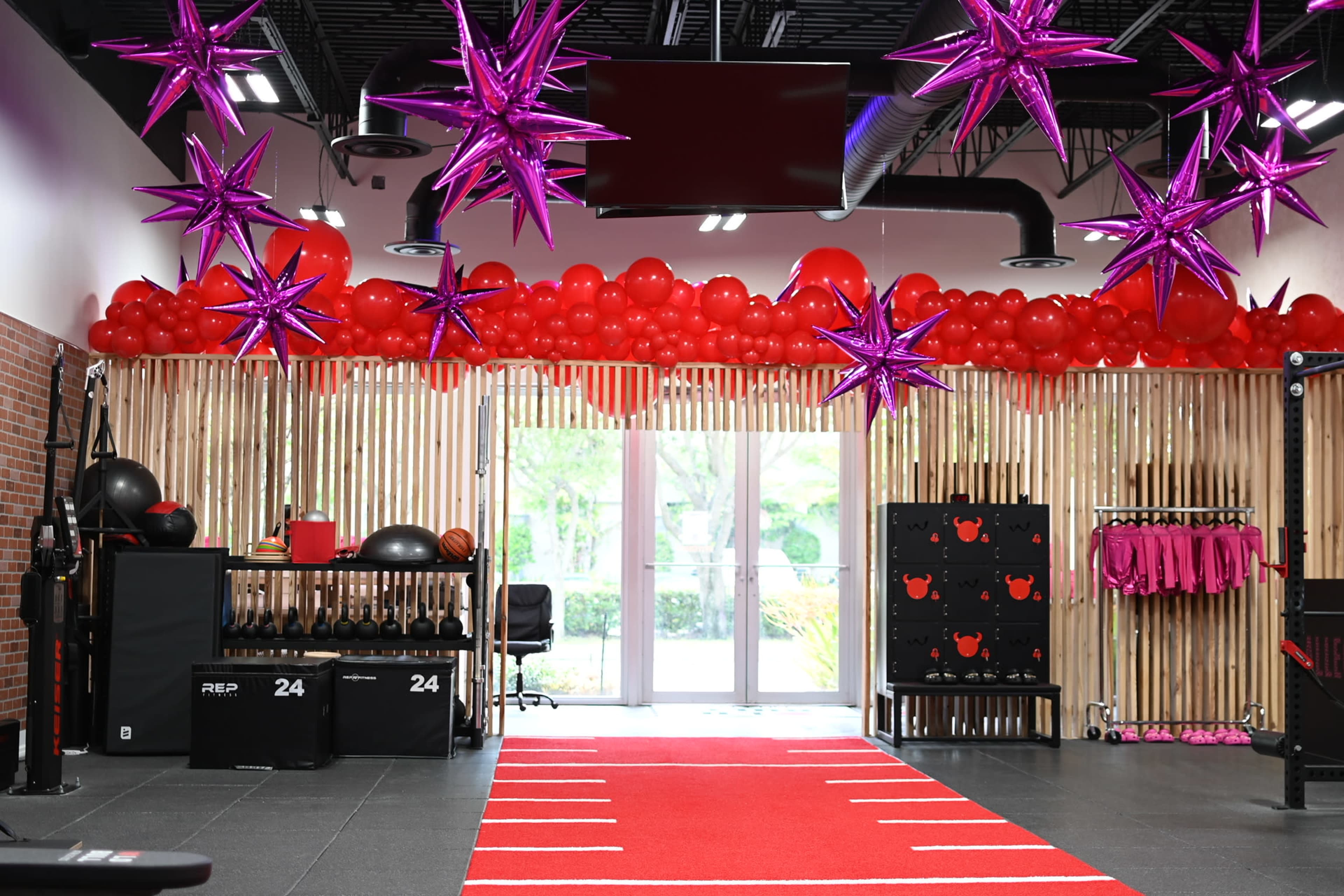 A fitness studio decorated with red balloons and purple star-shaped decorations, featuring exercise equipment and a red carpet leading to a glass door.