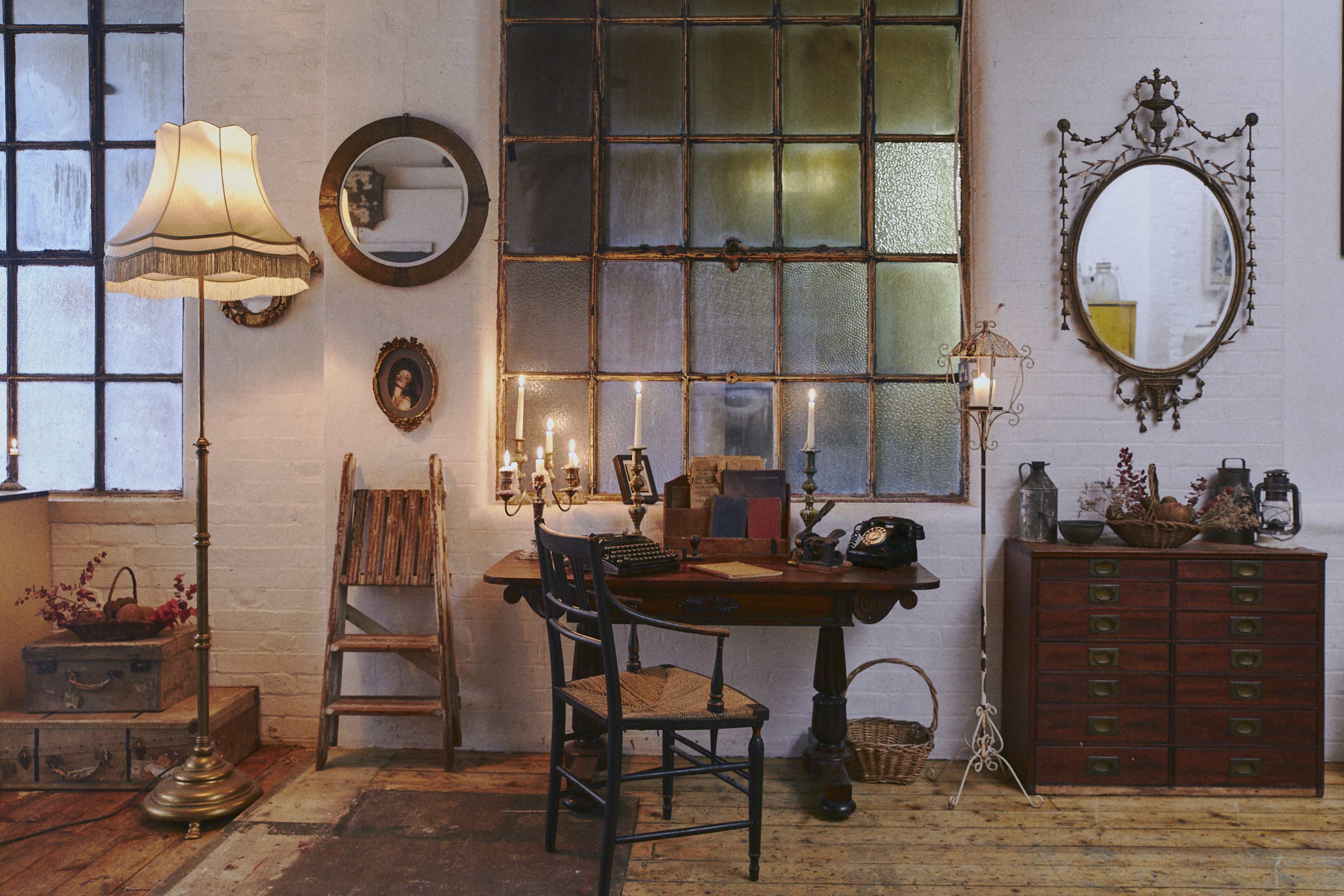 A vintage-inspired room featuring a wooden desk with a chair, a patterned lamp, mirrors, and various decorative items, all arranged near large windows.