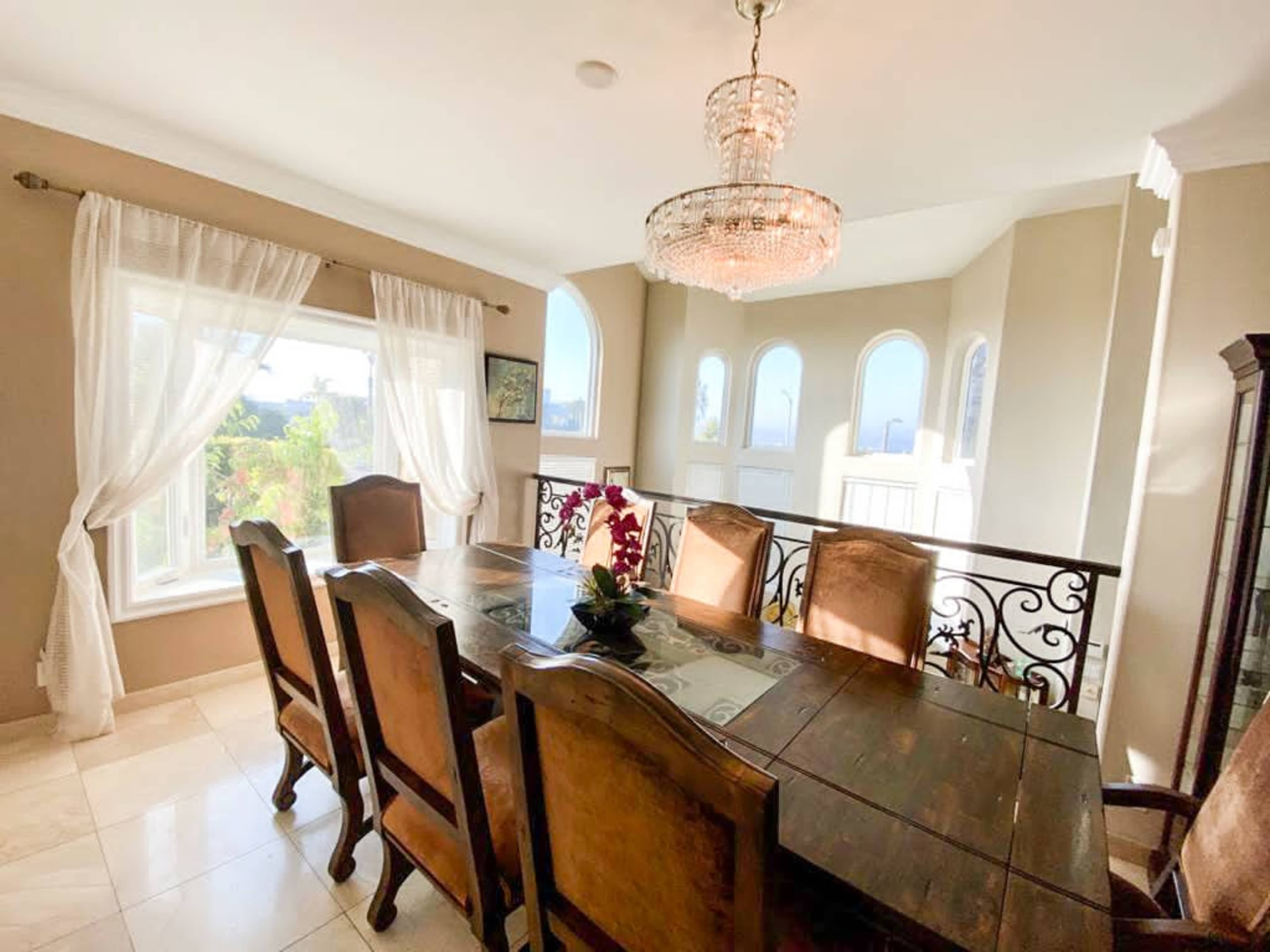 A wooden dining table surrounded by eight chairs is set in a room with large windows and a chandelier overhead.
