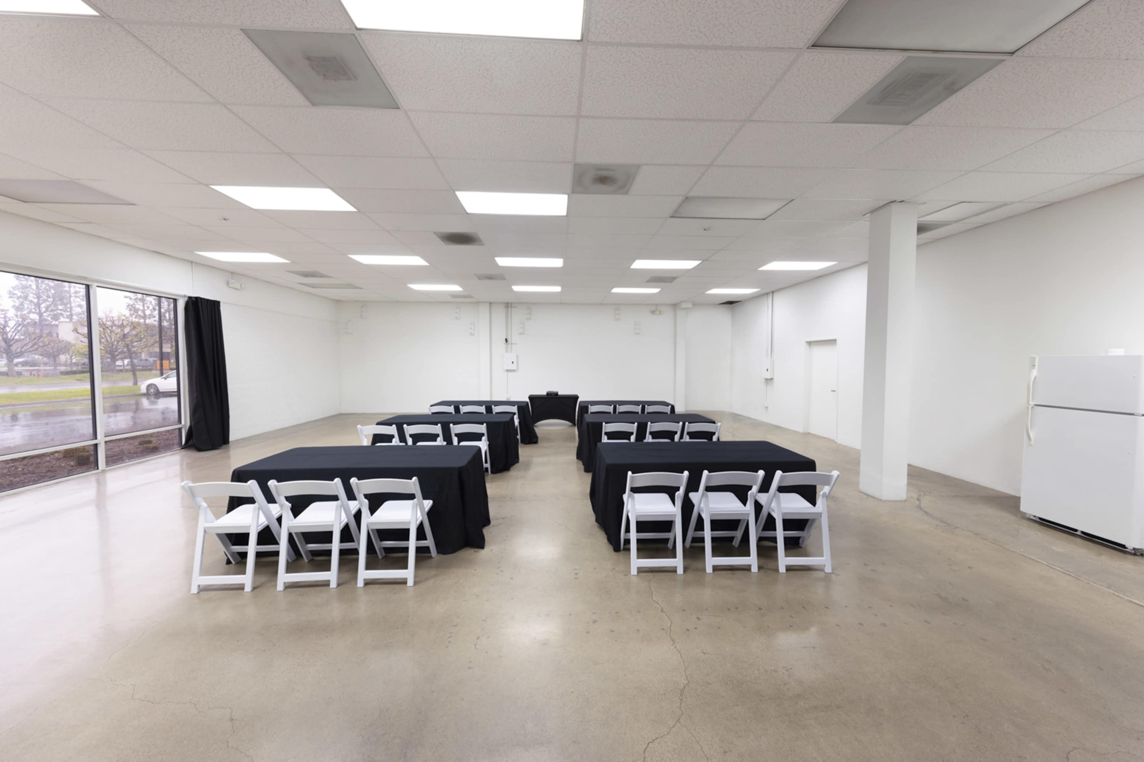 An empty event space features rows of tables covered with black tablecloths and surrounded by white folding chairs, with a refrigerator against one wall.