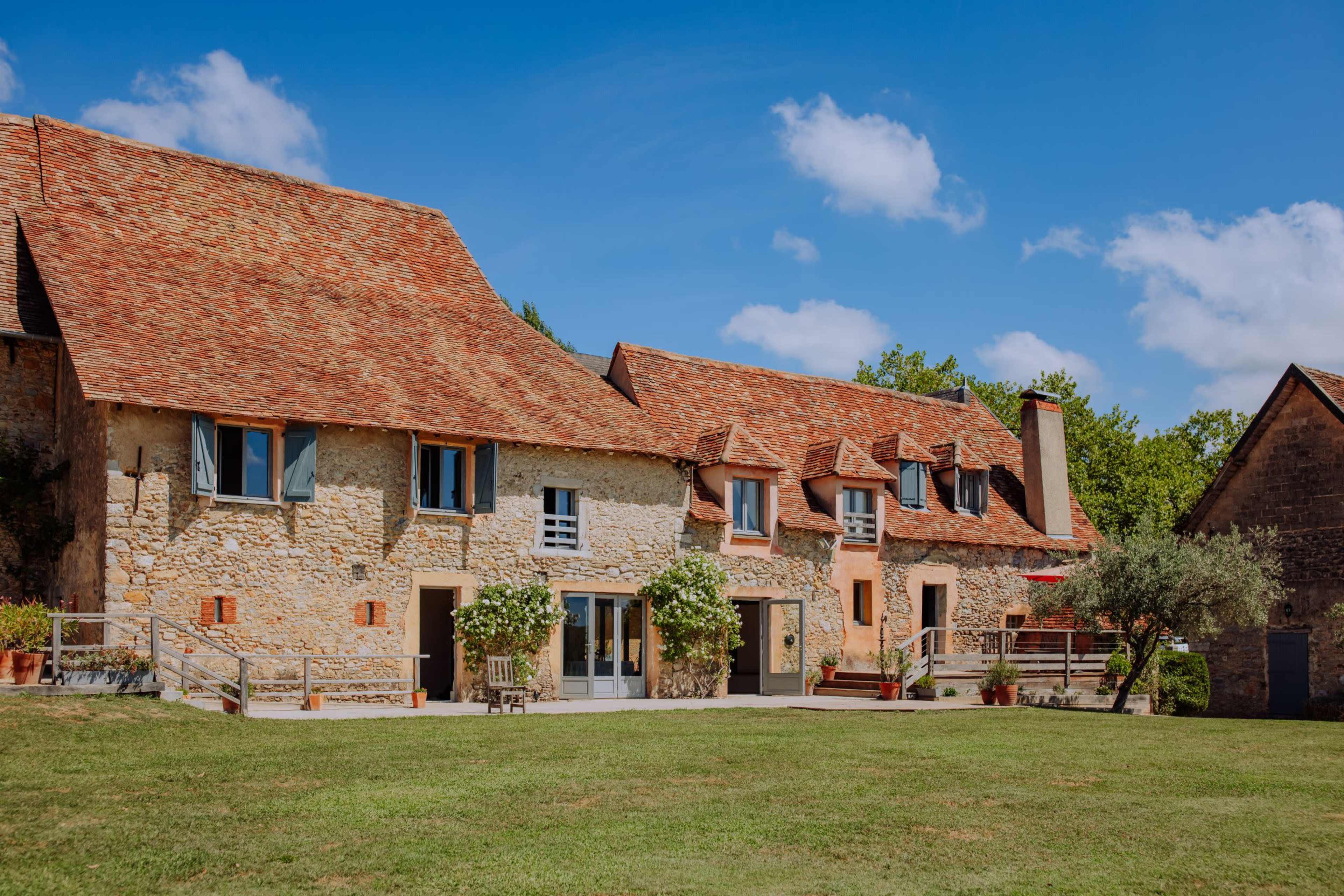 A stone house with a red-tiled roof and a grassy lawn is set against a blue sky with scattered clouds.