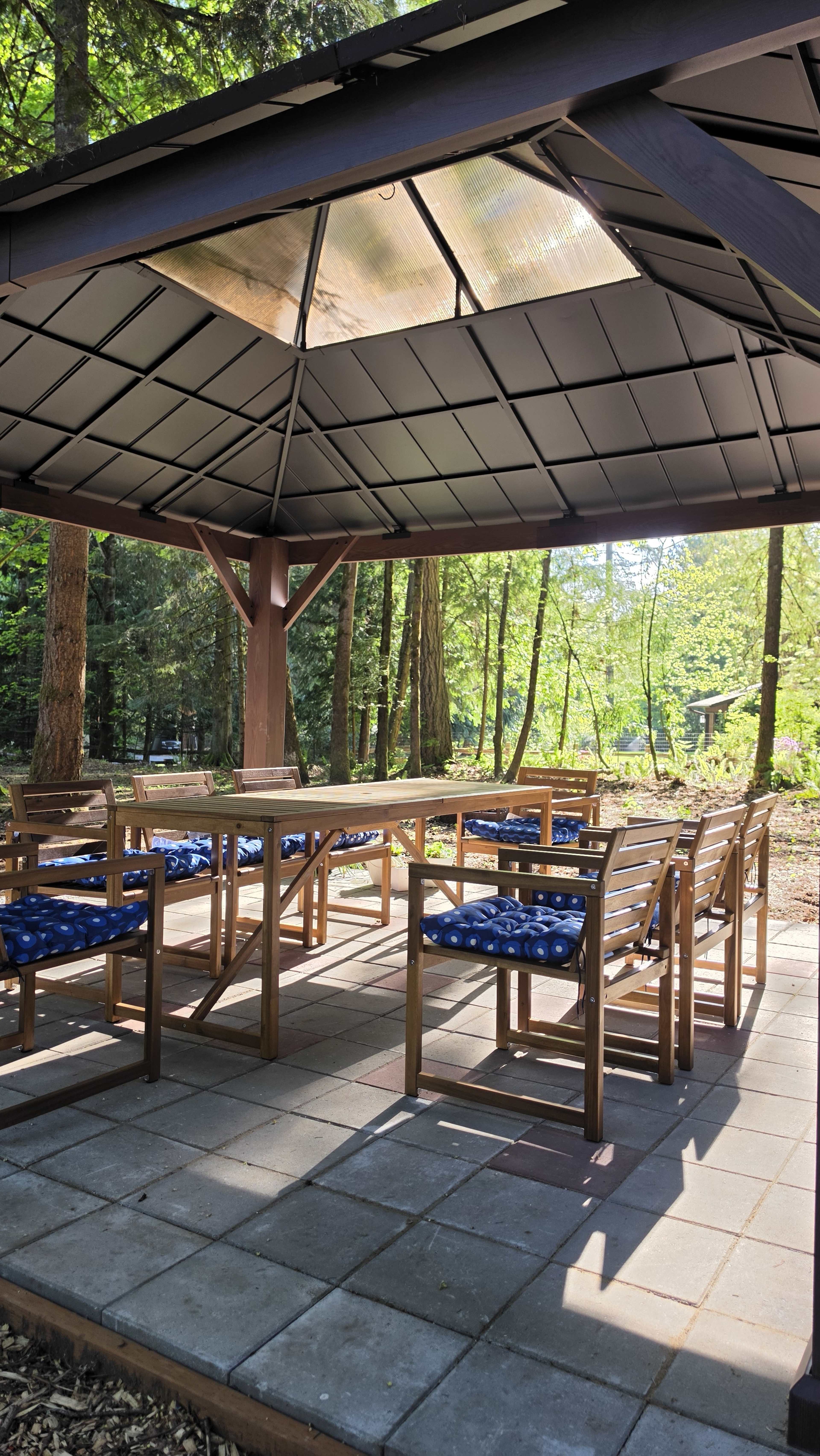 The image shows a gazebo with wooden seating and a table, surrounded by trees in a forested area.