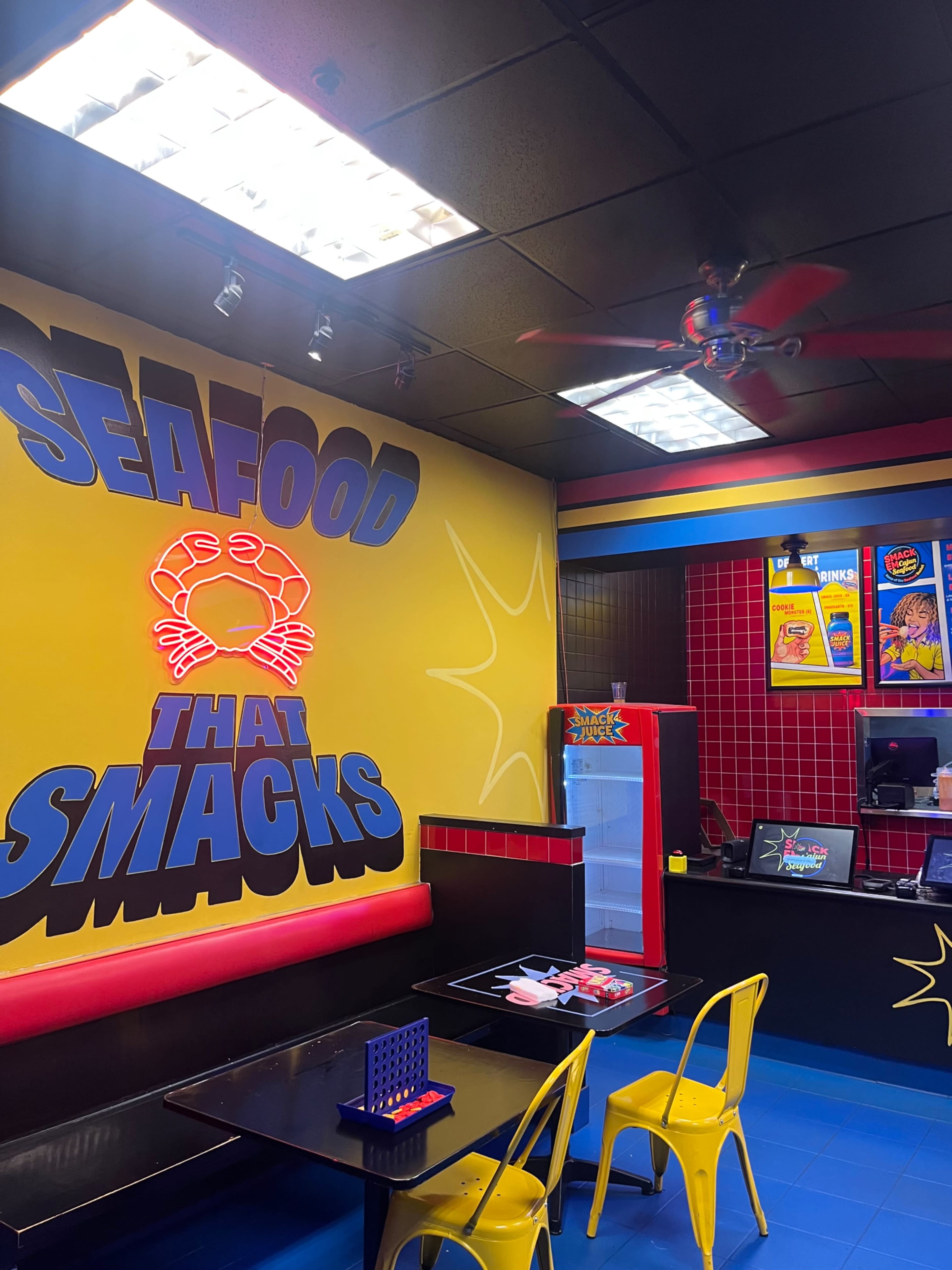 The interior of a colorful seafood restaurant features bright yellow walls with the phrase "SEAFOOD THAT SMACKS," black tables, and yellow chairs.