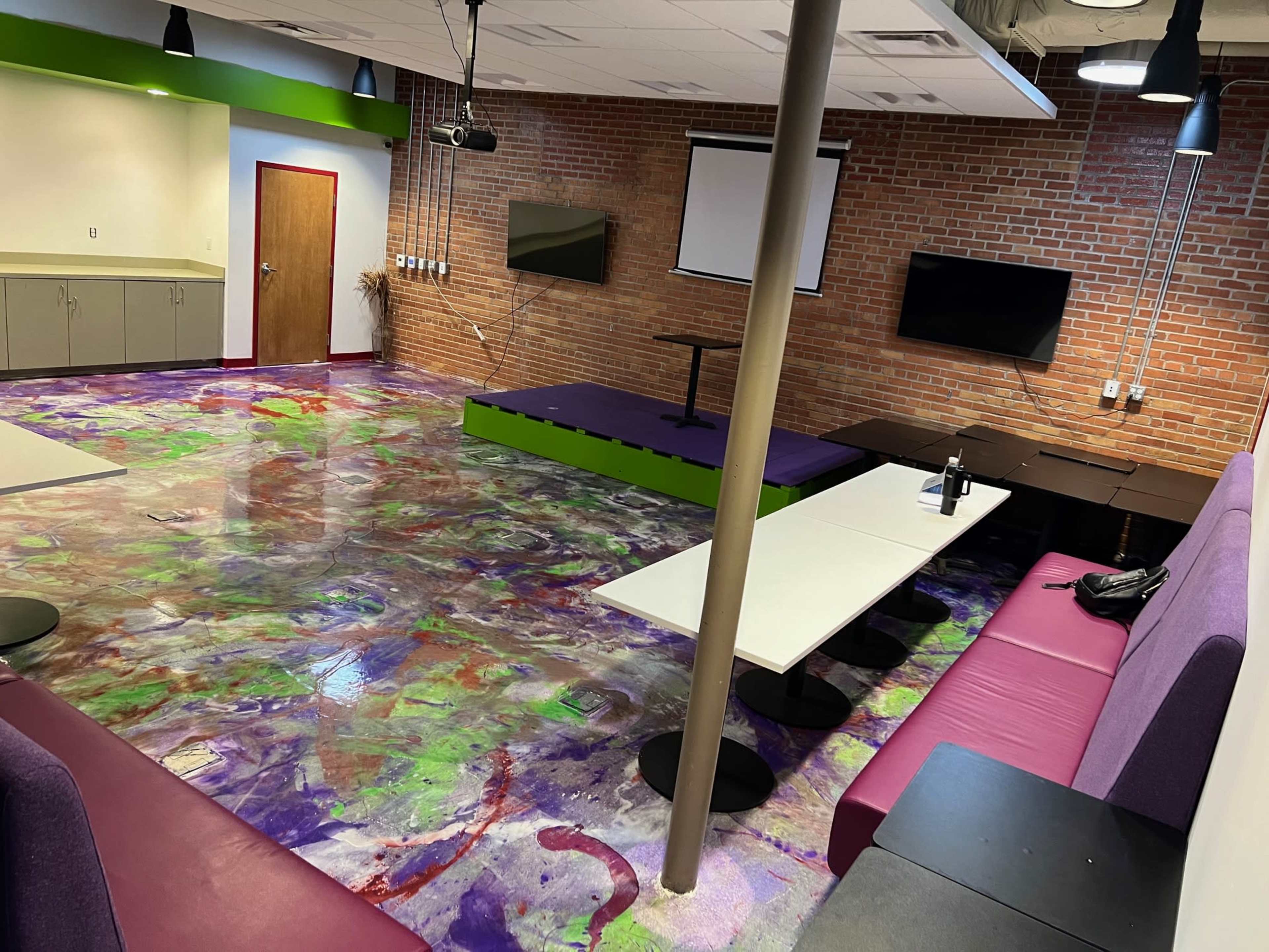 A vibrant, modern room with a colorful floor, exposed brick walls, seating areas, and a central table.