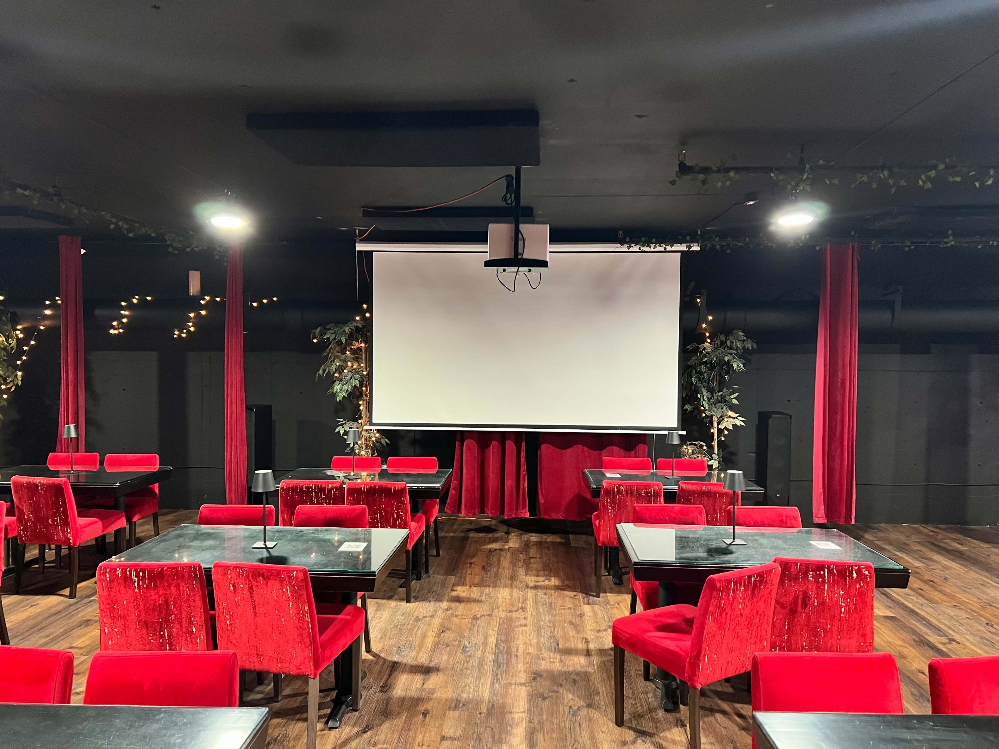 The image shows a dimly lit venue with red velvet seats, black tables, and a large white projection screen at the front.