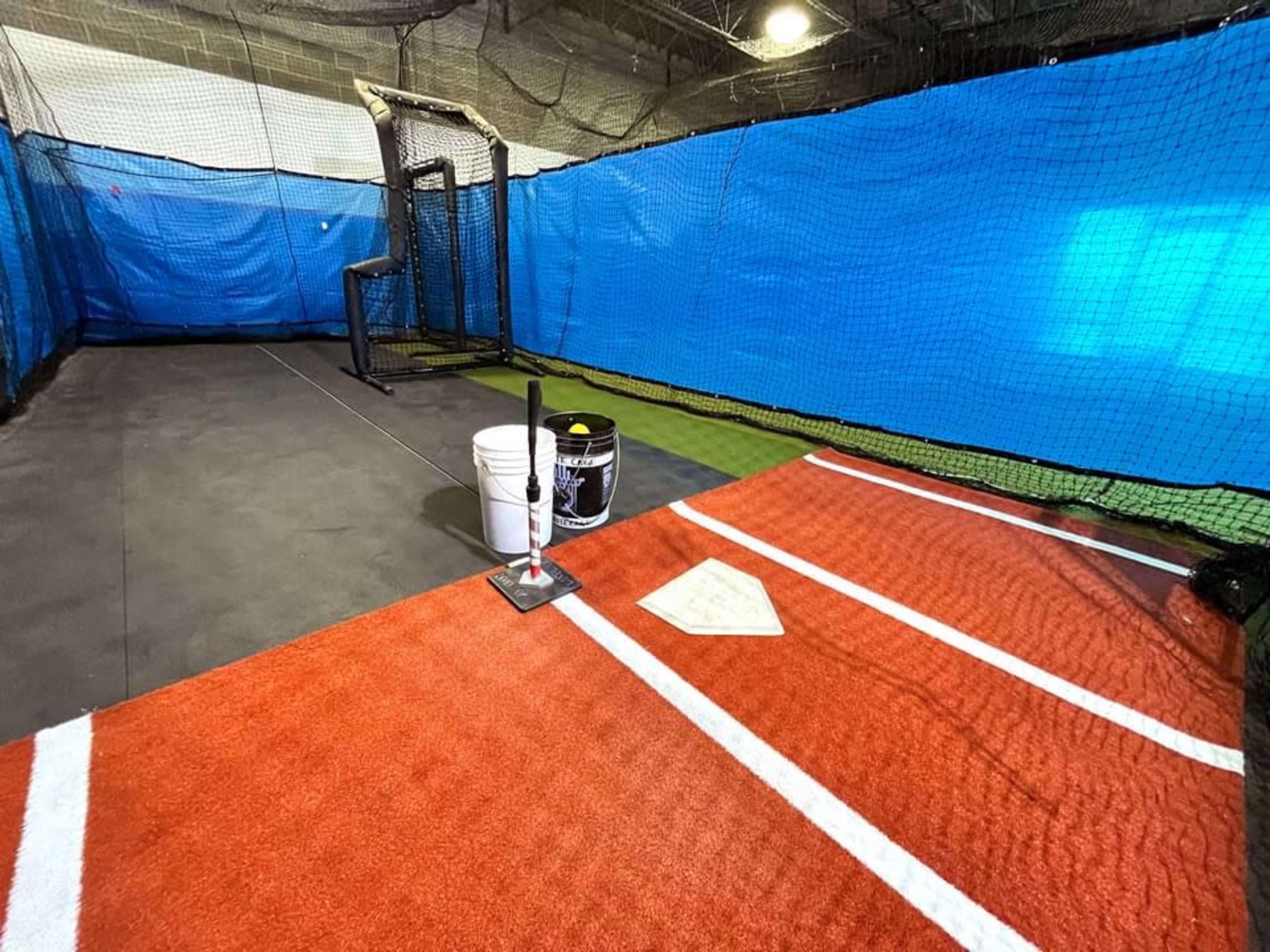 An indoor baseball batting cage with a red turf area, white home plate, and two buckets nearby.