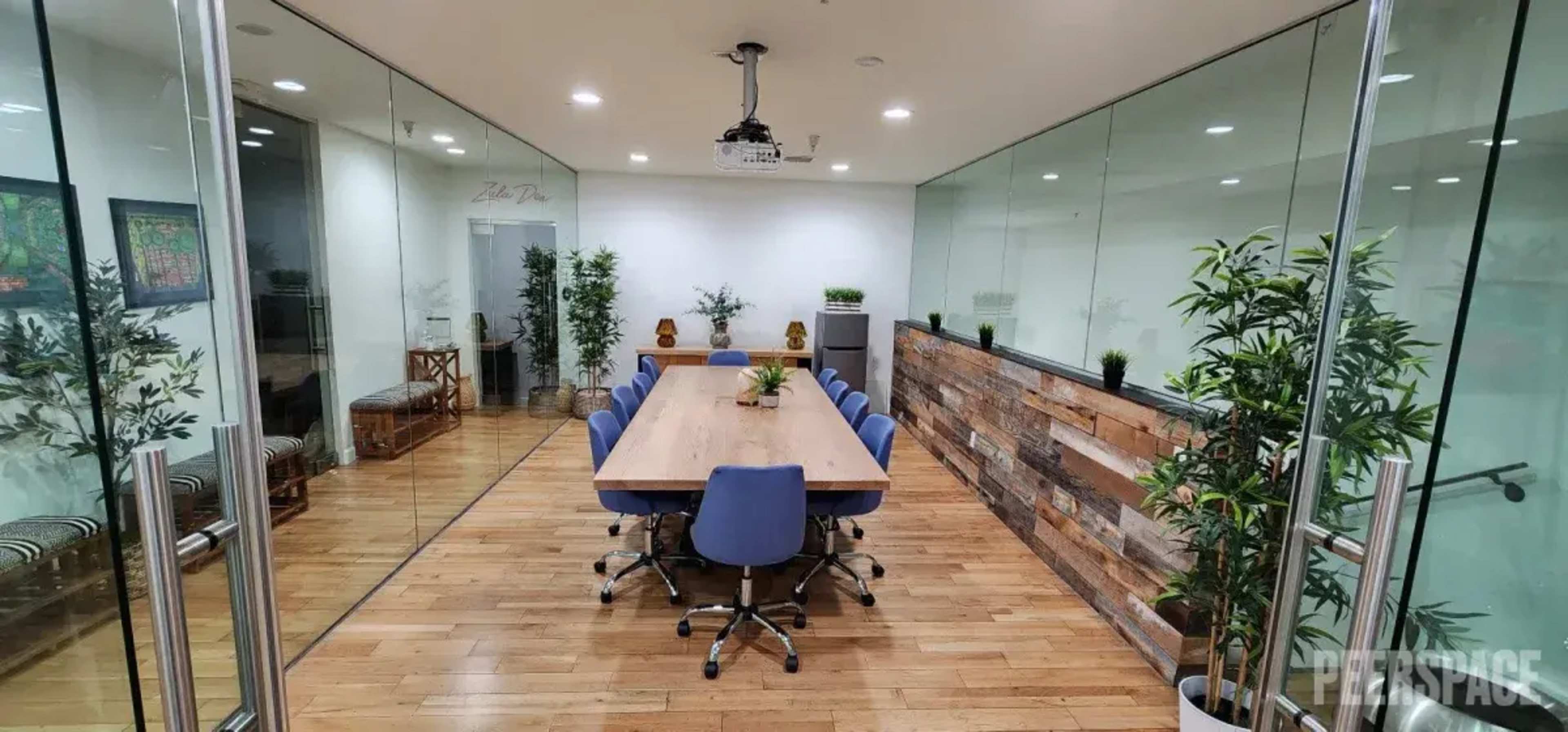 A modern conference room features a long wooden table surrounded by blue chairs, with glass walls and plants lining the space.