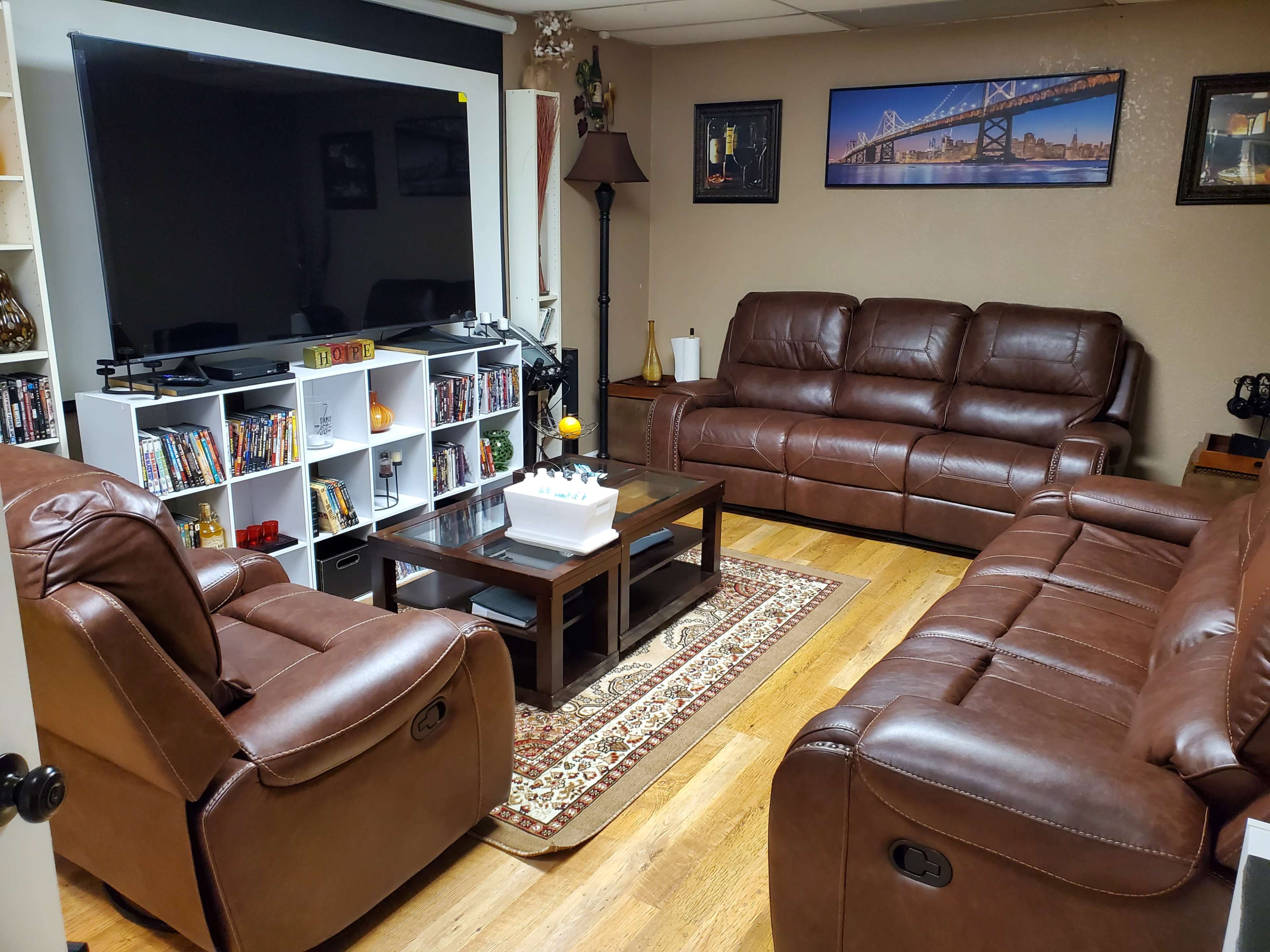 A living room features two brown leather couches facing a coffee table, with a large television mounted on the wall and a bookshelf filled with DVDs in the corner.