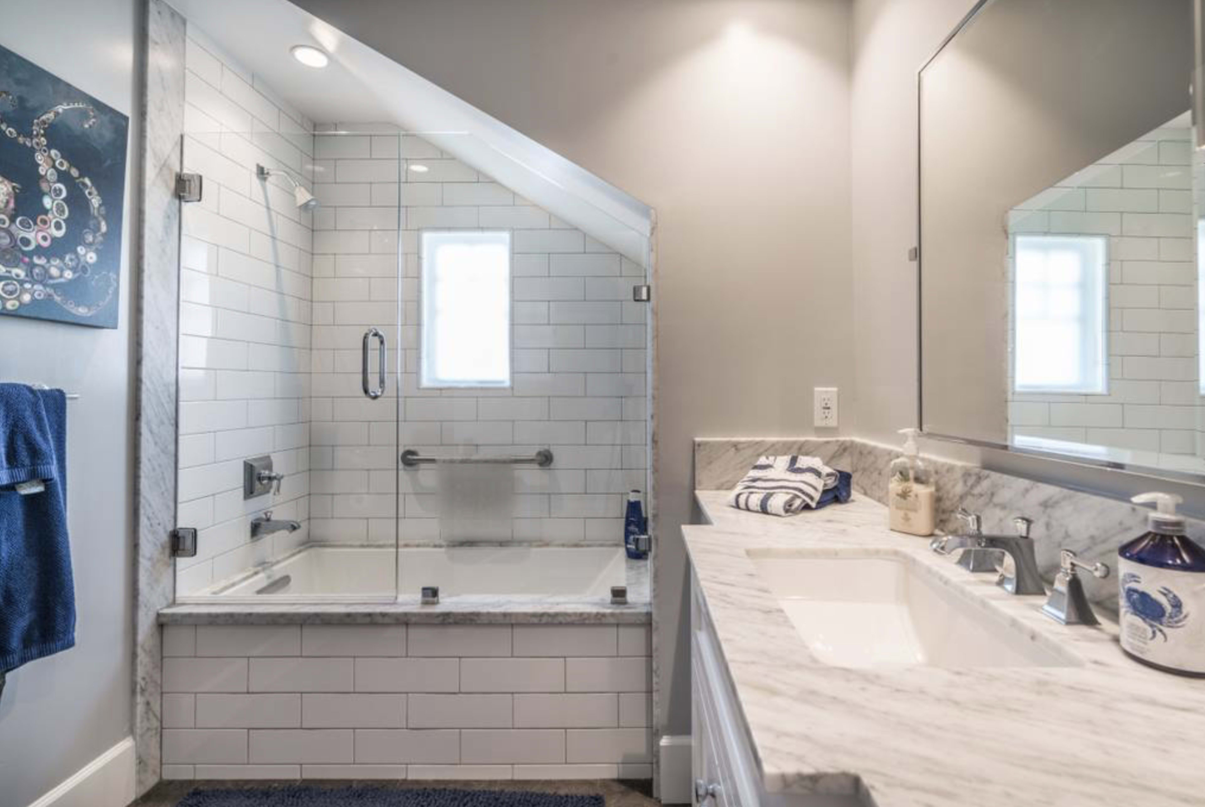 The image shows a modern bathroom with a glass-enclosed shower, a white bathtub, and a marble countertop with two sinks.