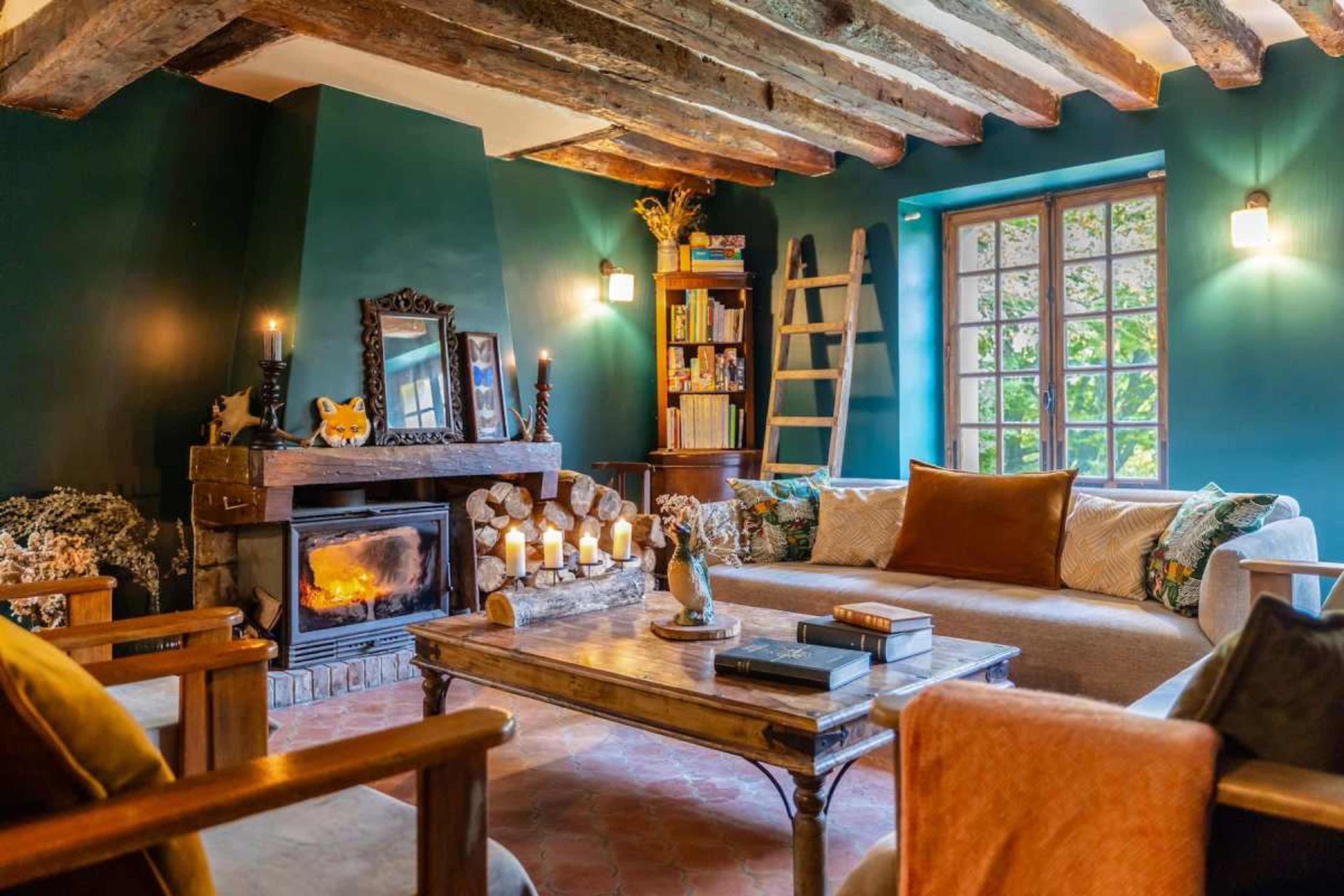 The image depicts a cozy living room featuring a stone fireplace, wooden beams on the ceiling, and a mix of comfortable seating and shelves filled with books.