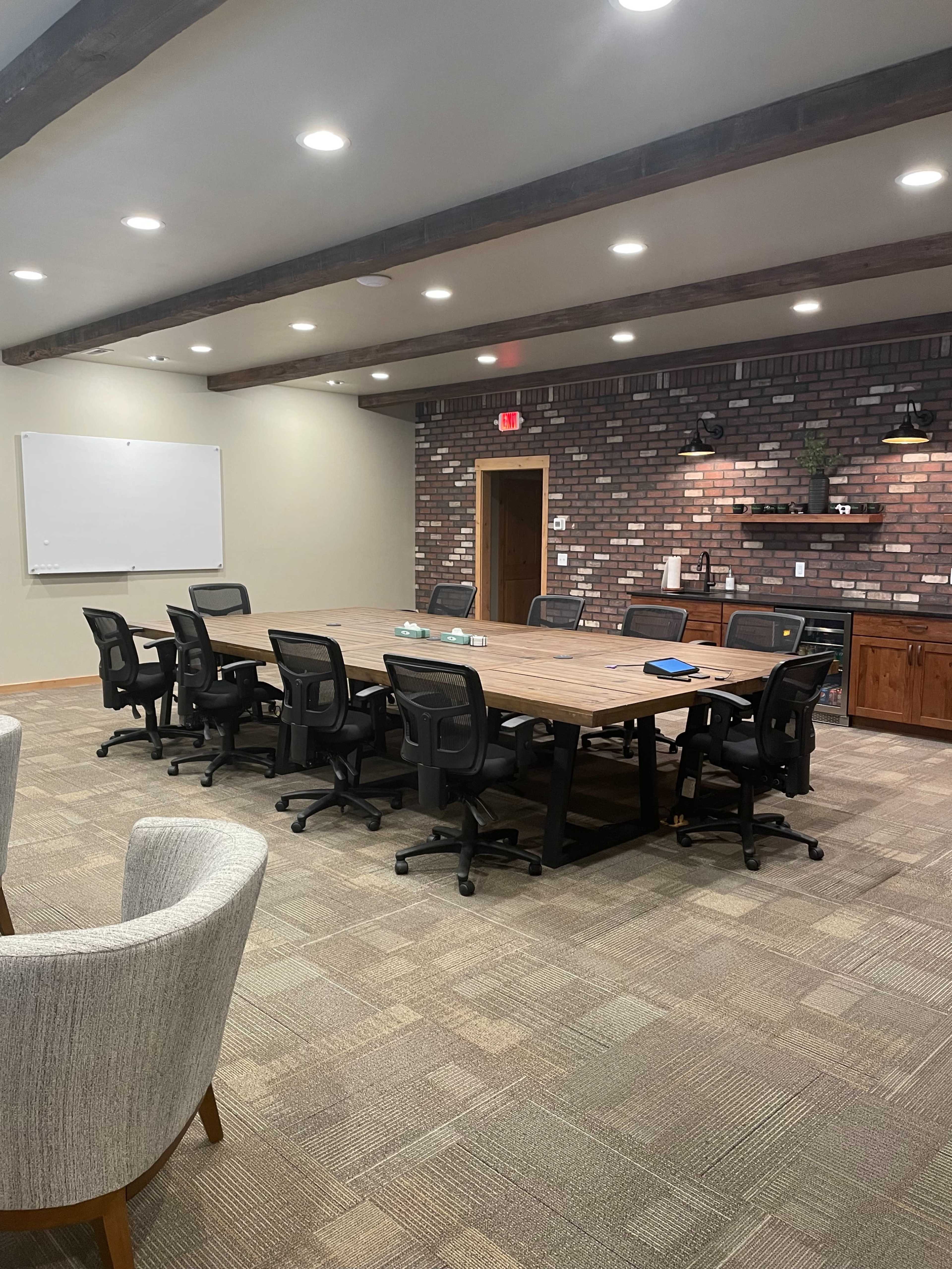 A spacious conference room features a large wooden table surrounded by office chairs, with a brick wall and cabinets in the background.