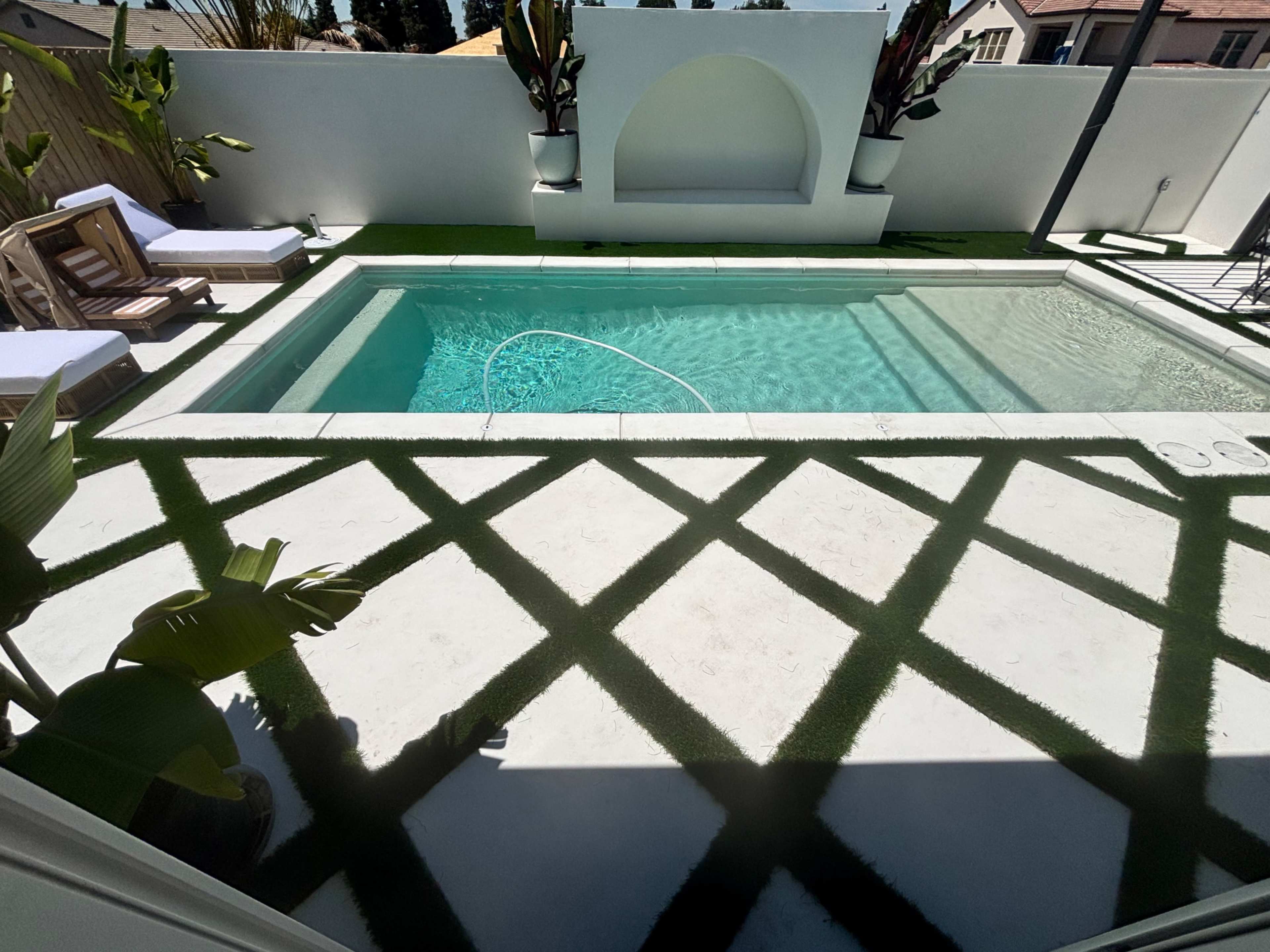 The image shows a swimming pool surrounded by a white wall, sleek lounge chairs, and green artificial grass creating a modern outdoor space.