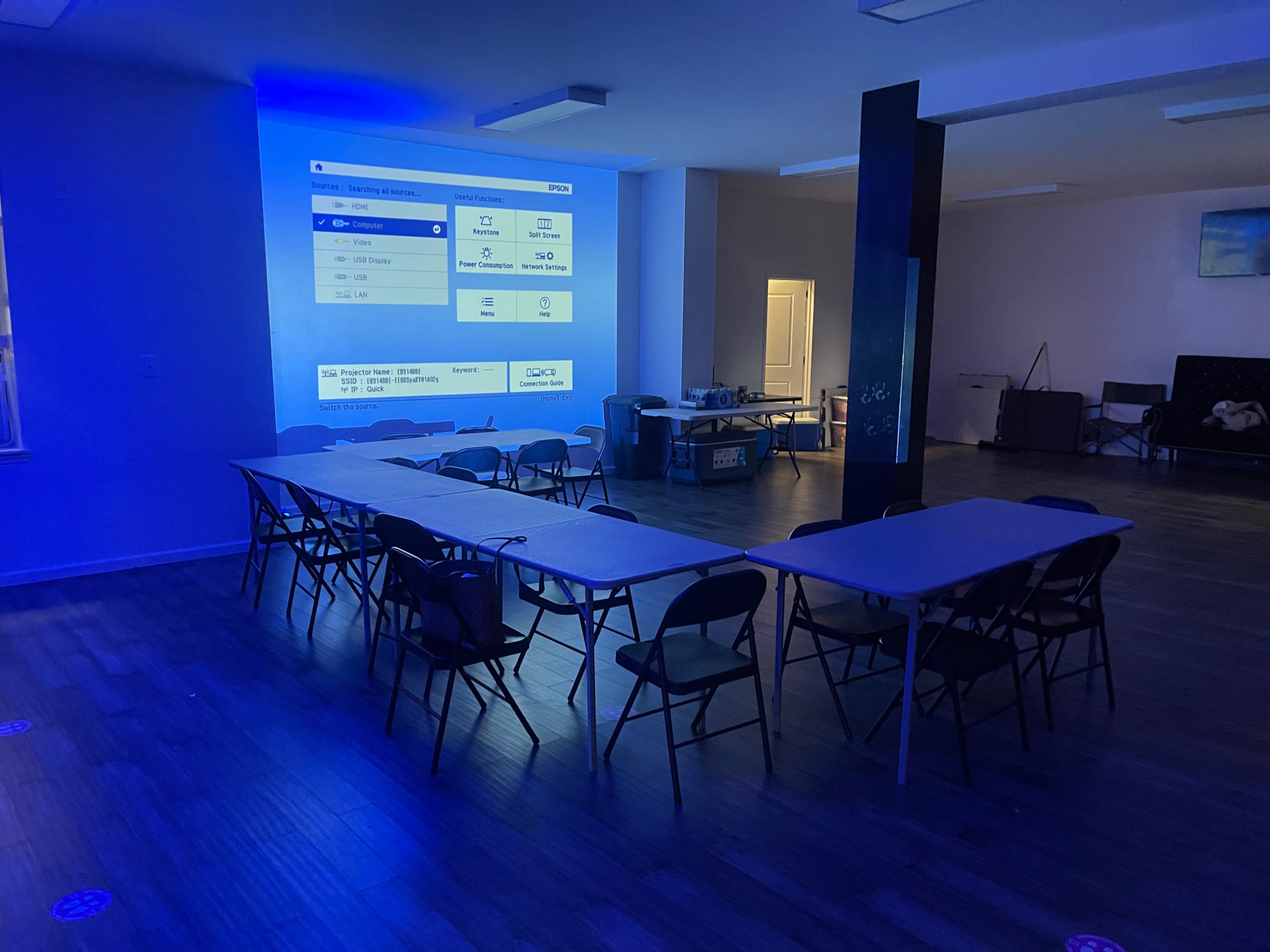 A room with blue lighting features a long row of folding tables and chairs arranged towards a wall displaying a large screen with digital content.
