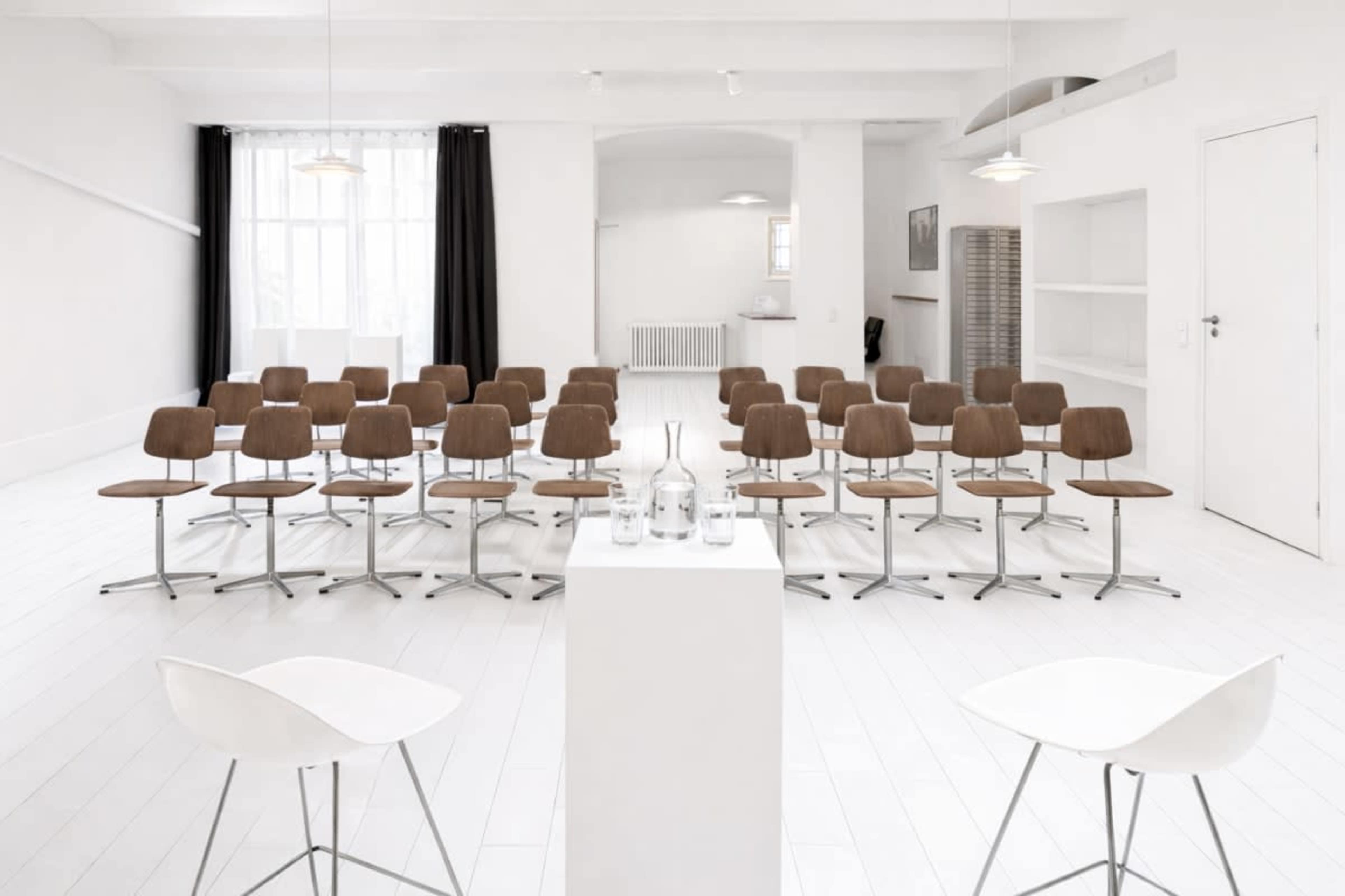 A minimalistic room features rows of wooden chairs facing a small white table with glass bottles, illuminated by natural light.