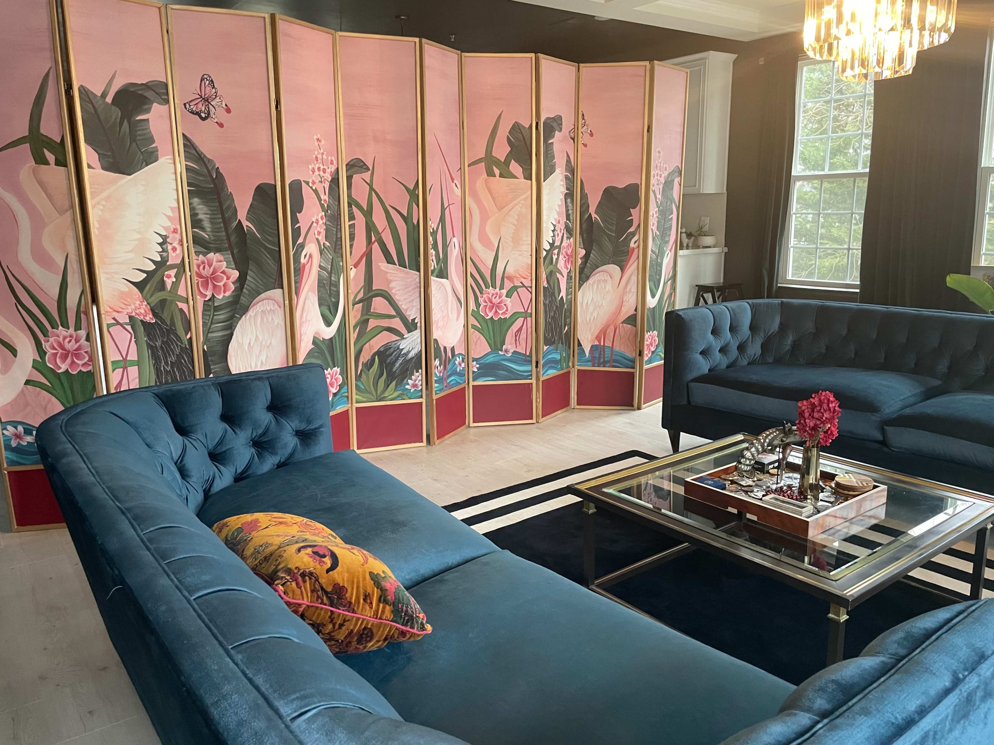 A living room features a blue velvet sectional sofa, a glass coffee table with decorative items, and a pink folding screen adorned with cranes and tropical plants.