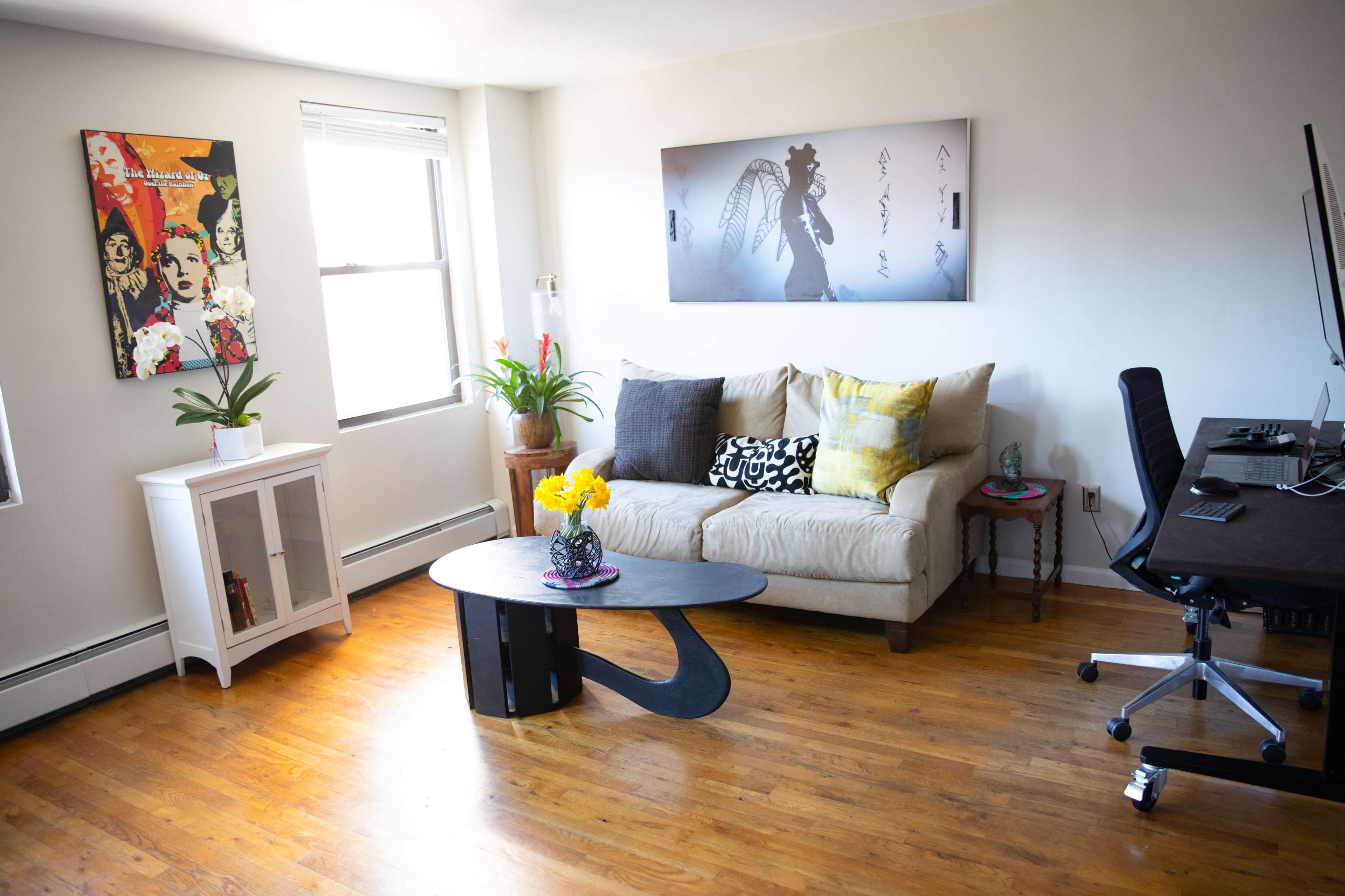 The image shows a modern living room with a sofa, a coffee table, a small cabinet, and an adjacent workspace featuring a desk and computer.