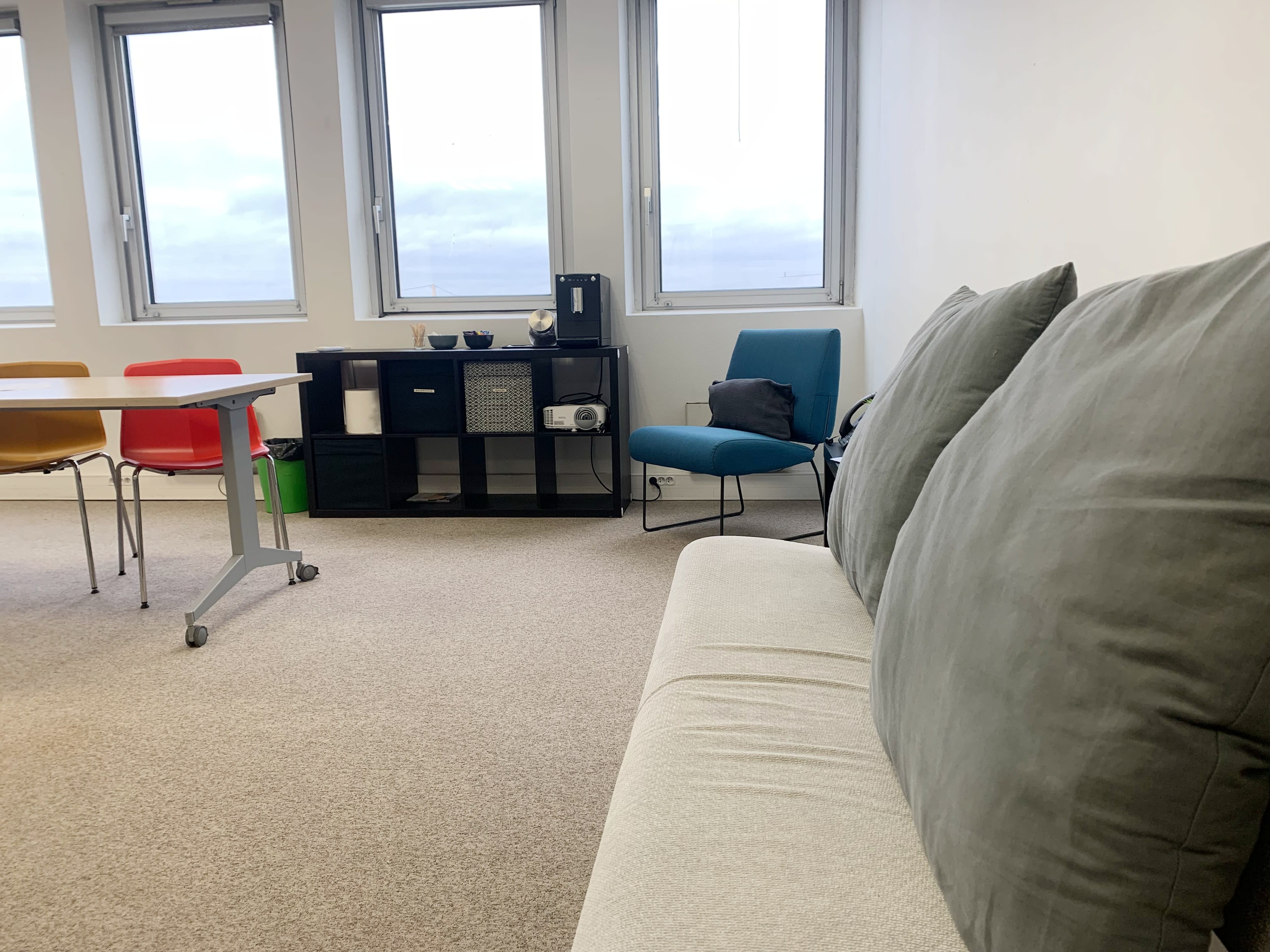A well-lit room with large windows, a grey sofa, a small table with chairs, and a storage unit displaying a coffee maker and other items.
