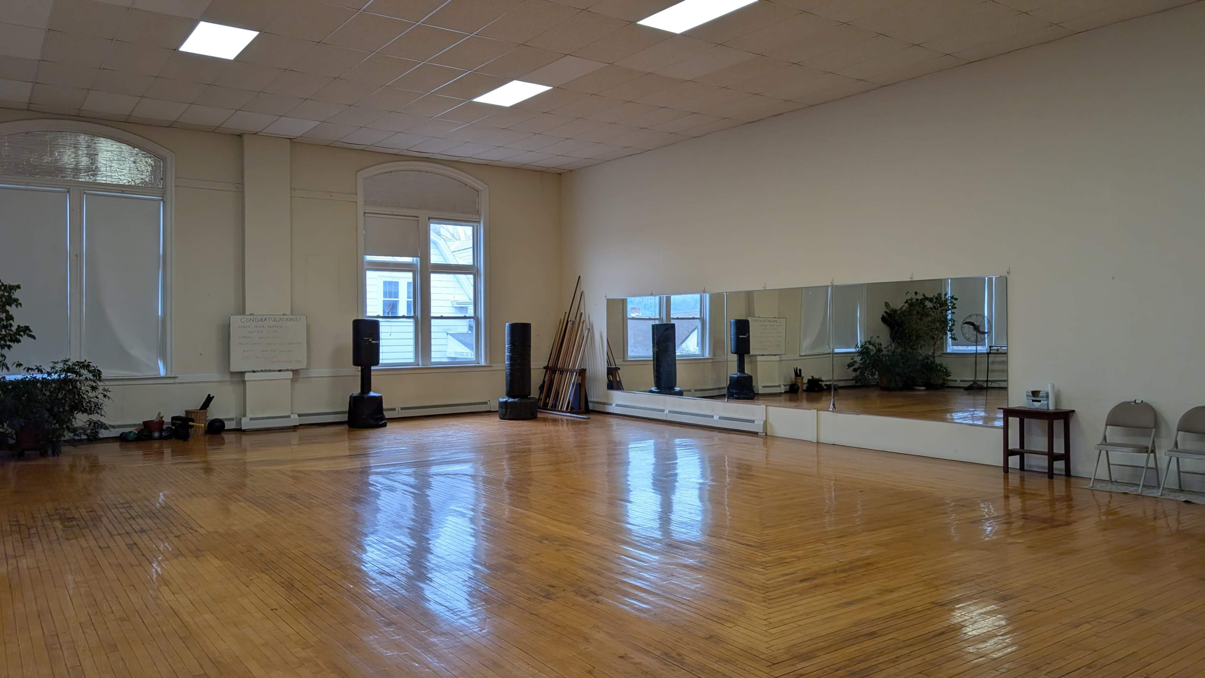 The image shows a spacious dance studio with wooden flooring, large mirrors, and natural light coming through windows.