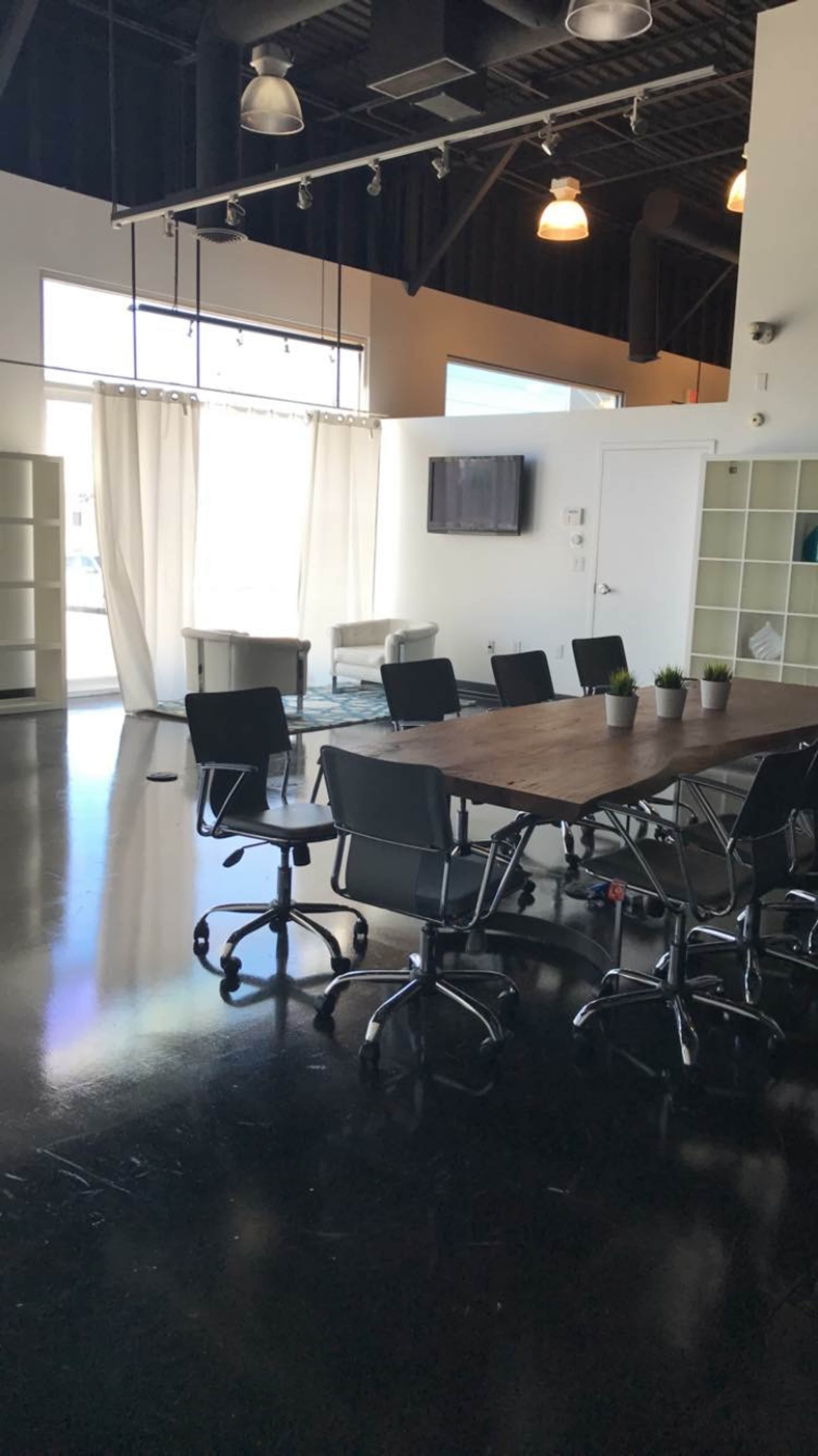 A modern conference room with a long wooden table surrounded by black chairs, a television mounted on the wall, and large windows letting in natural light.
