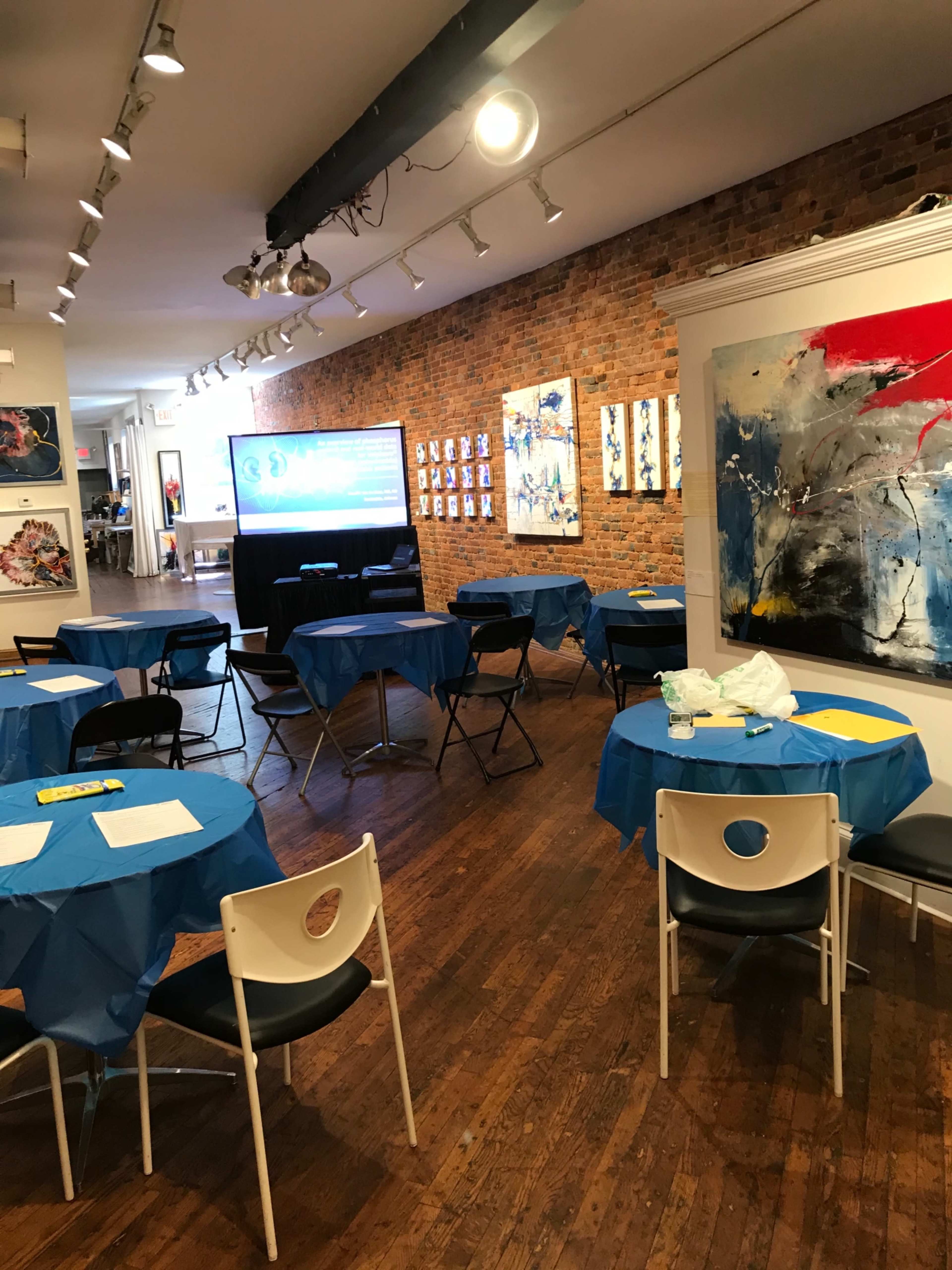A spacious gallery features several blue-covered tables and chairs, with artwork displayed on the walls and a screen at the front.