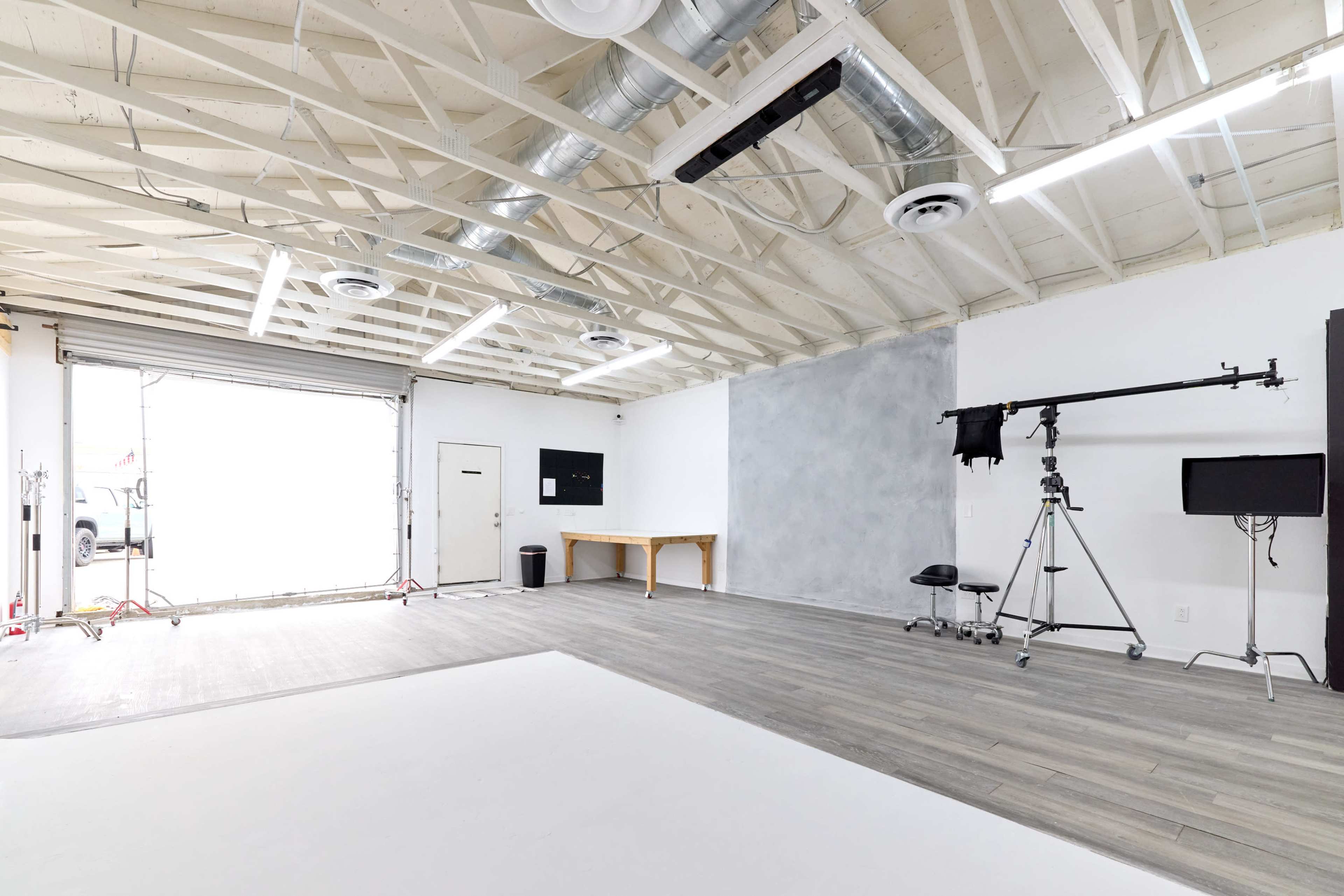 A spacious, empty studio with a light gray wall, a large window, a table, and equipment including a camera stand and monitors.