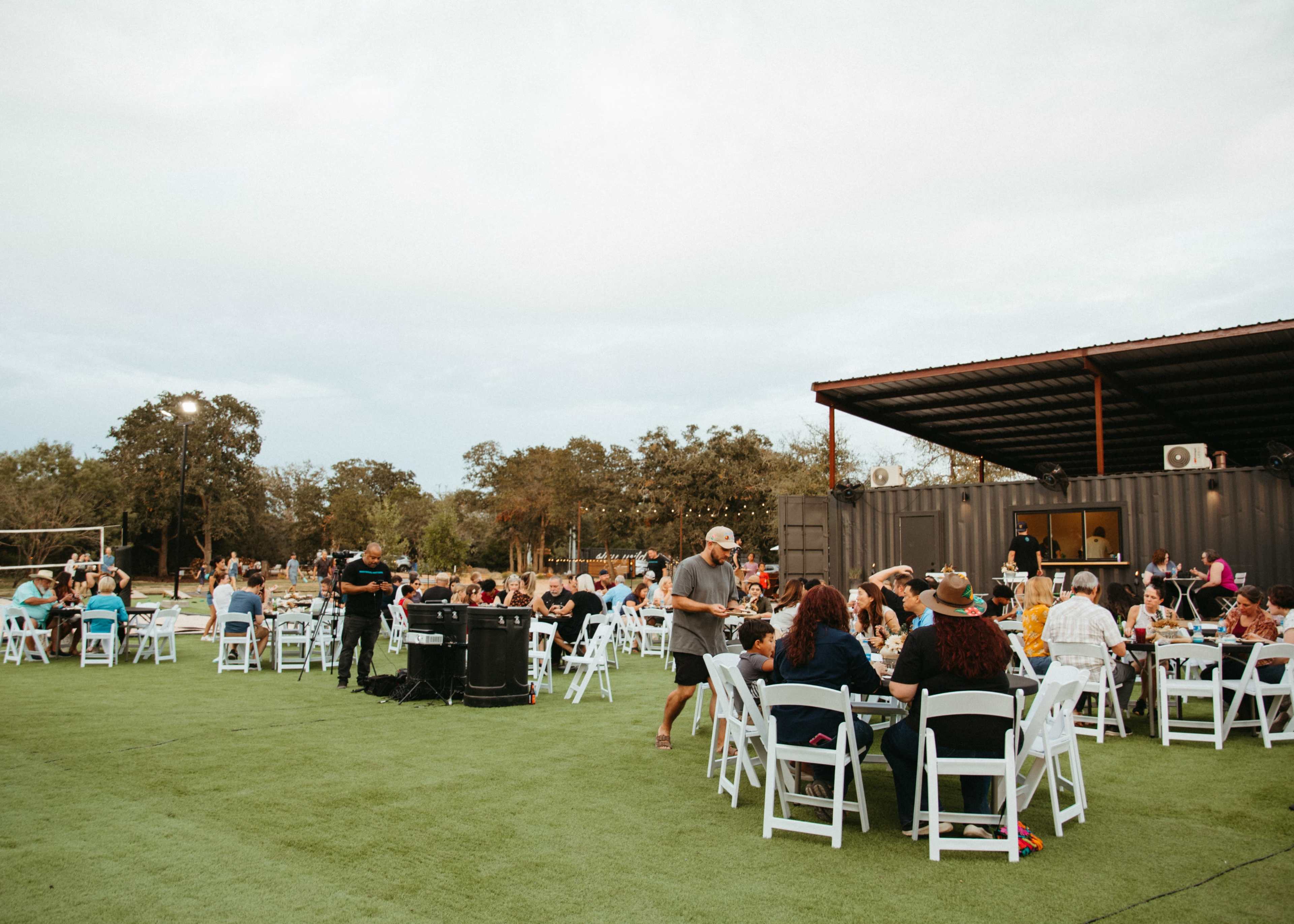 A large outdoor gathering features numerous tables filled with people, set up on green turf beneath a cloudy sky.