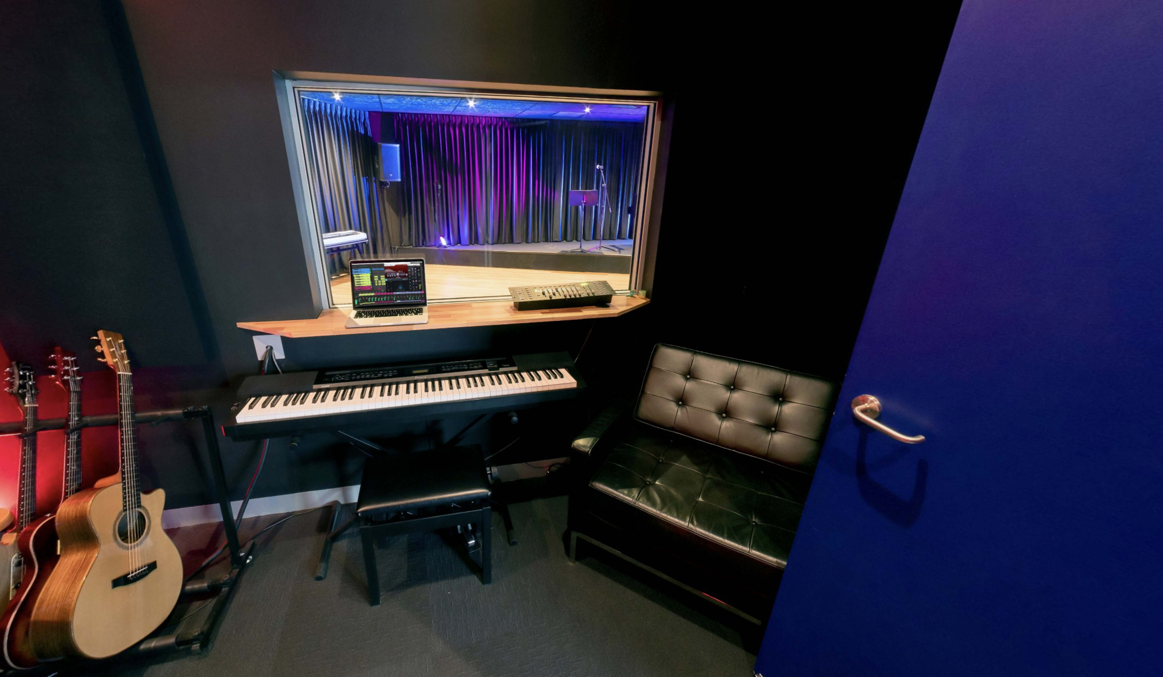 The image shows a small music studio corner featuring a keyboard, a computer on a desktop, a black leather chair, and acoustic guitars leaning against the wall.