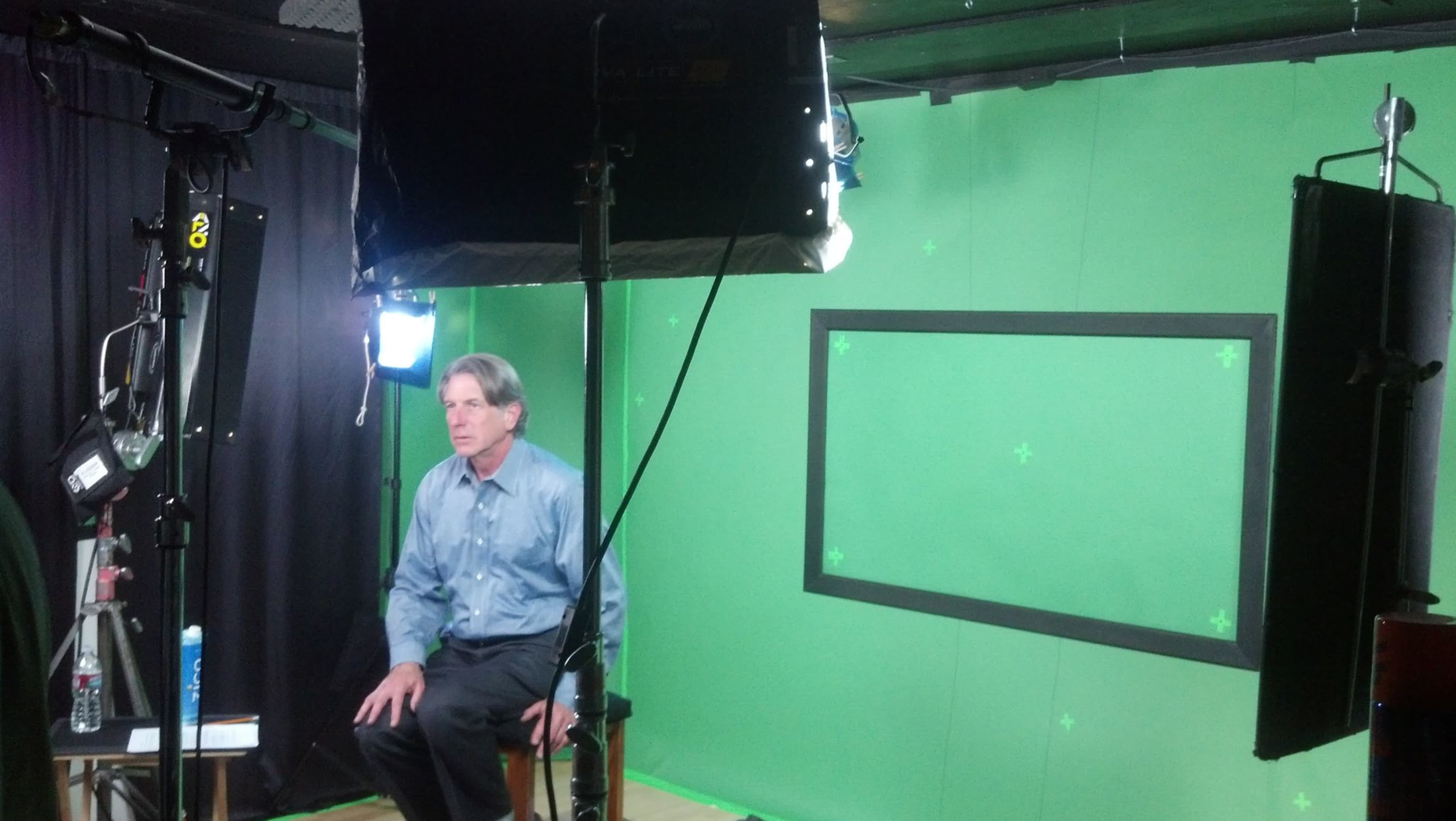 A man sits on a stool in front of a green-screen setup, with studio lights illuminating the scene.