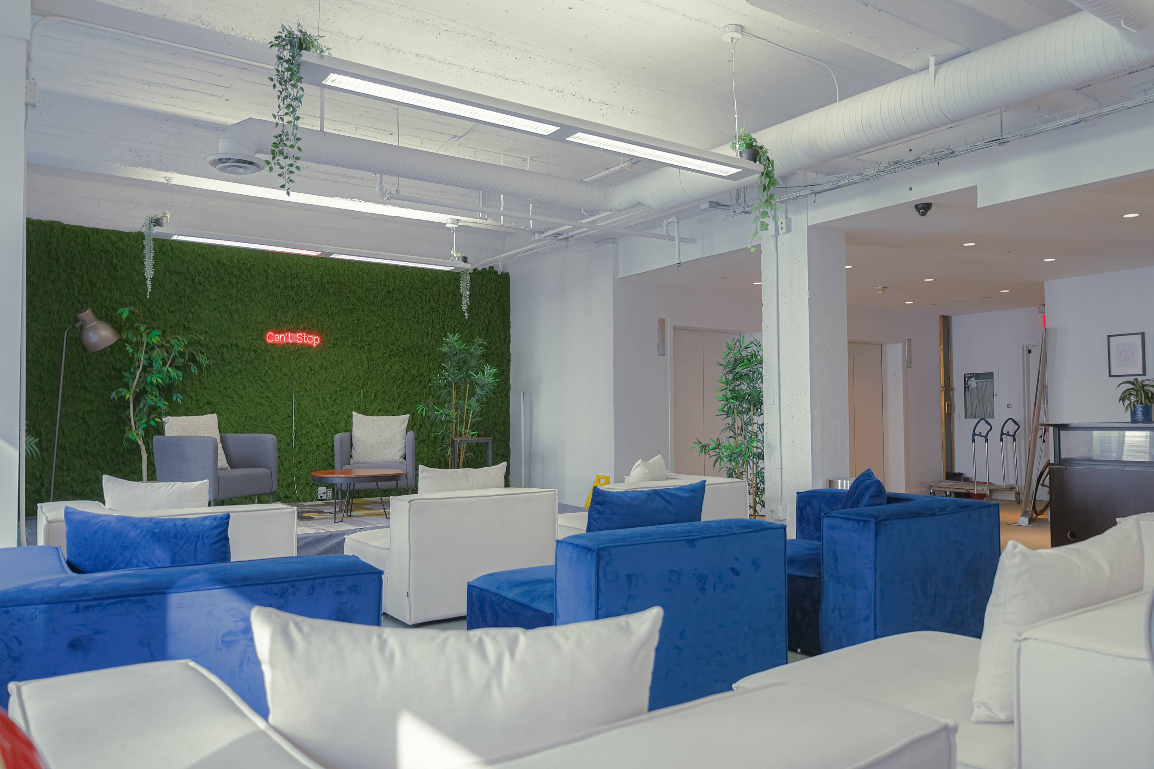 A modern lounge area features blue and white sofas, indoor plants, and a green wall with a neon sign.