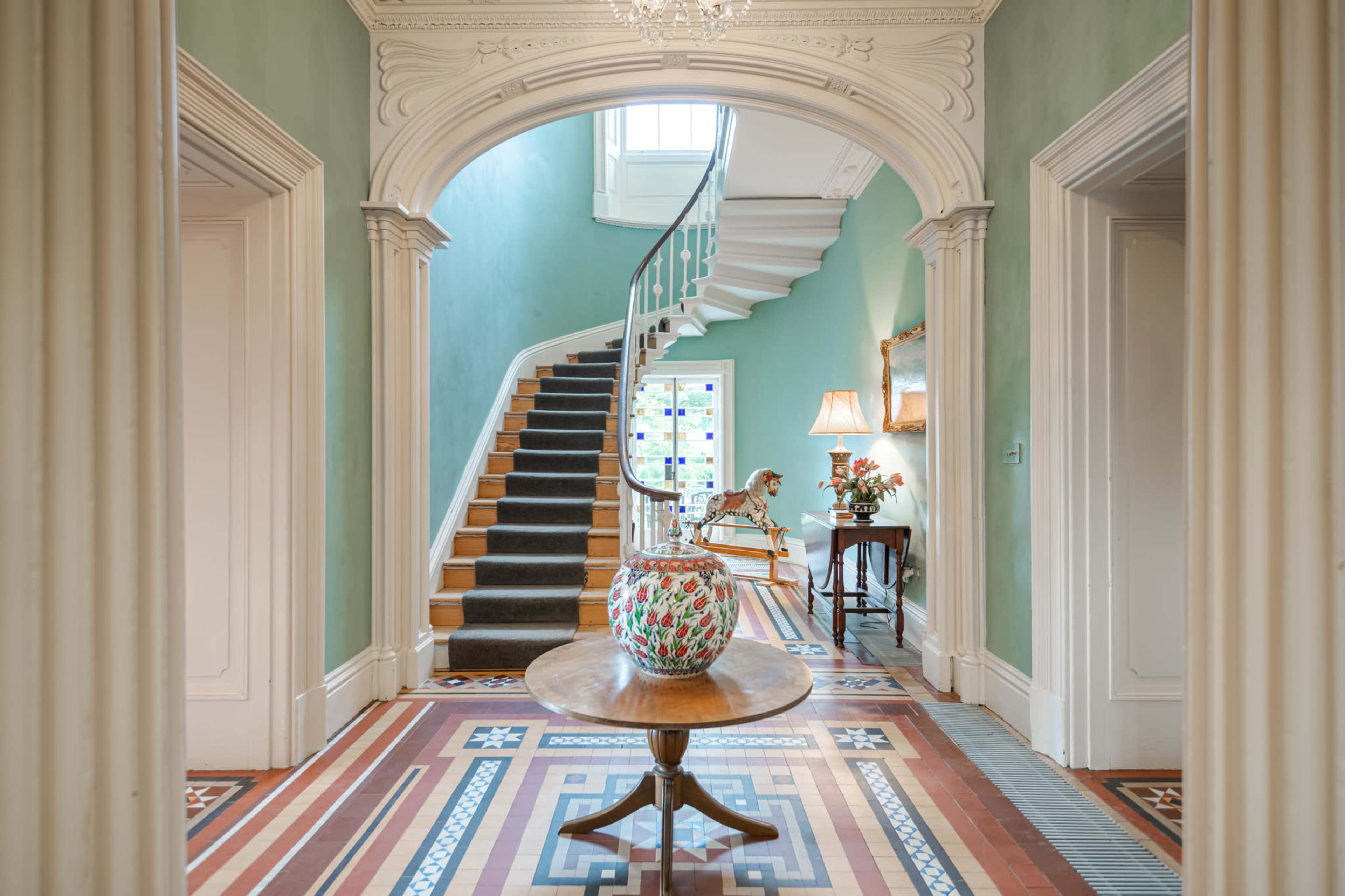 A spacious hallway features a curved staircase and a decorative table with a vase, flanked by two doorways and illuminated by natural light.