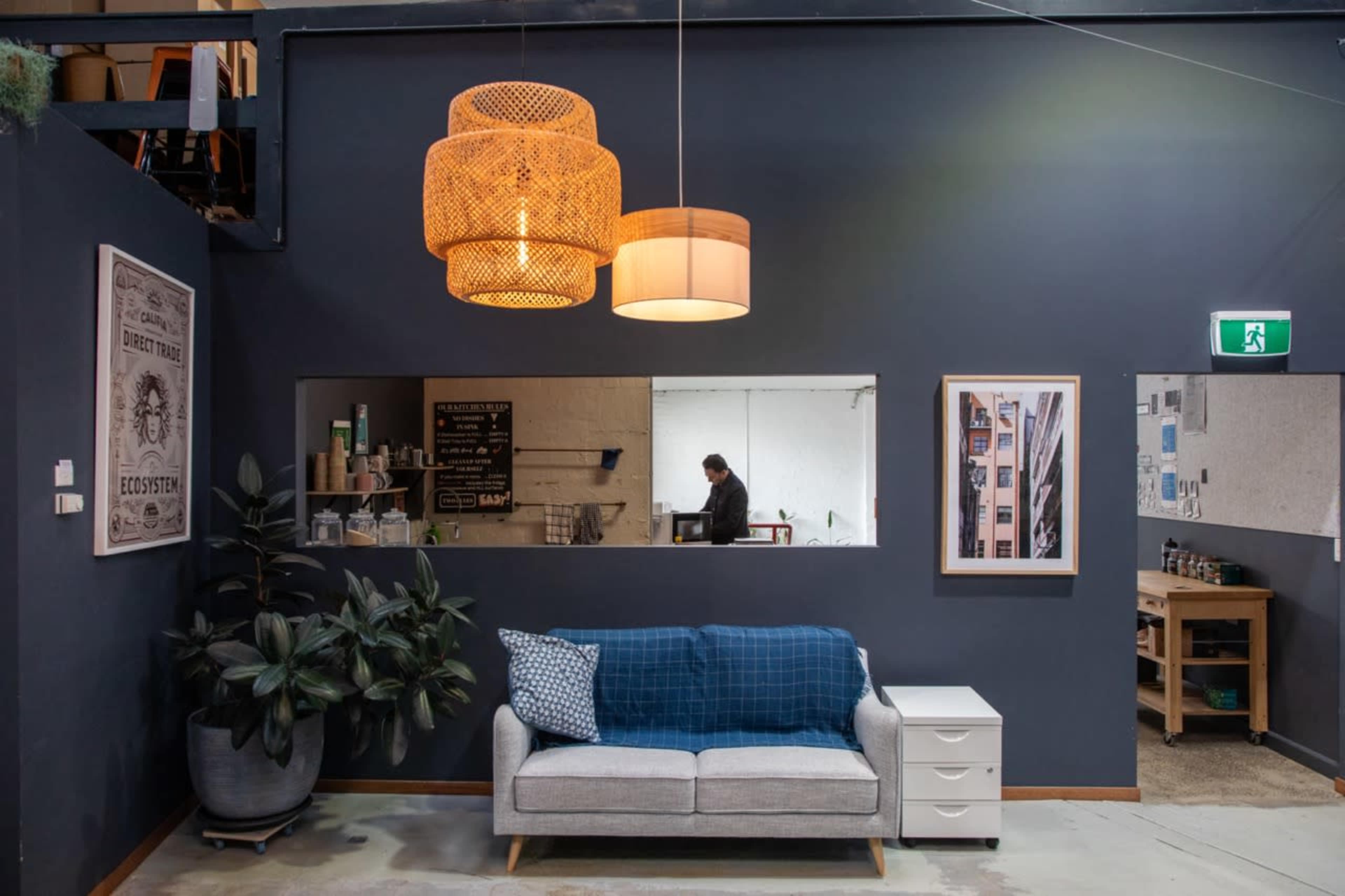 The image shows a modern indoor workspace featuring a sofa with a blue throw blanket, a large light fixture, and a kitchen area in the background.