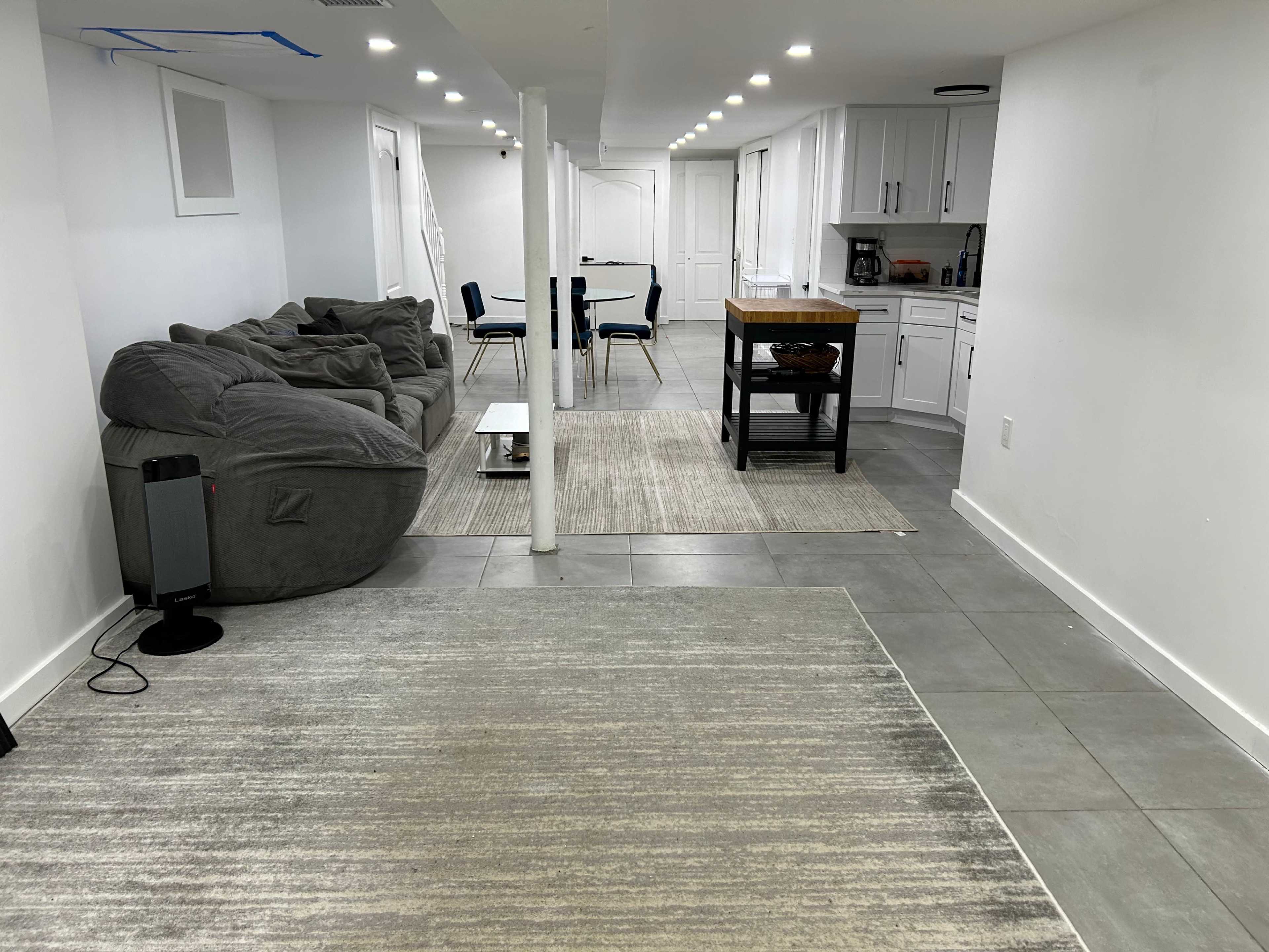 The image shows a spacious basement area featuring a gray sectional couch, a dining table with chairs, and a small kitchen island.
