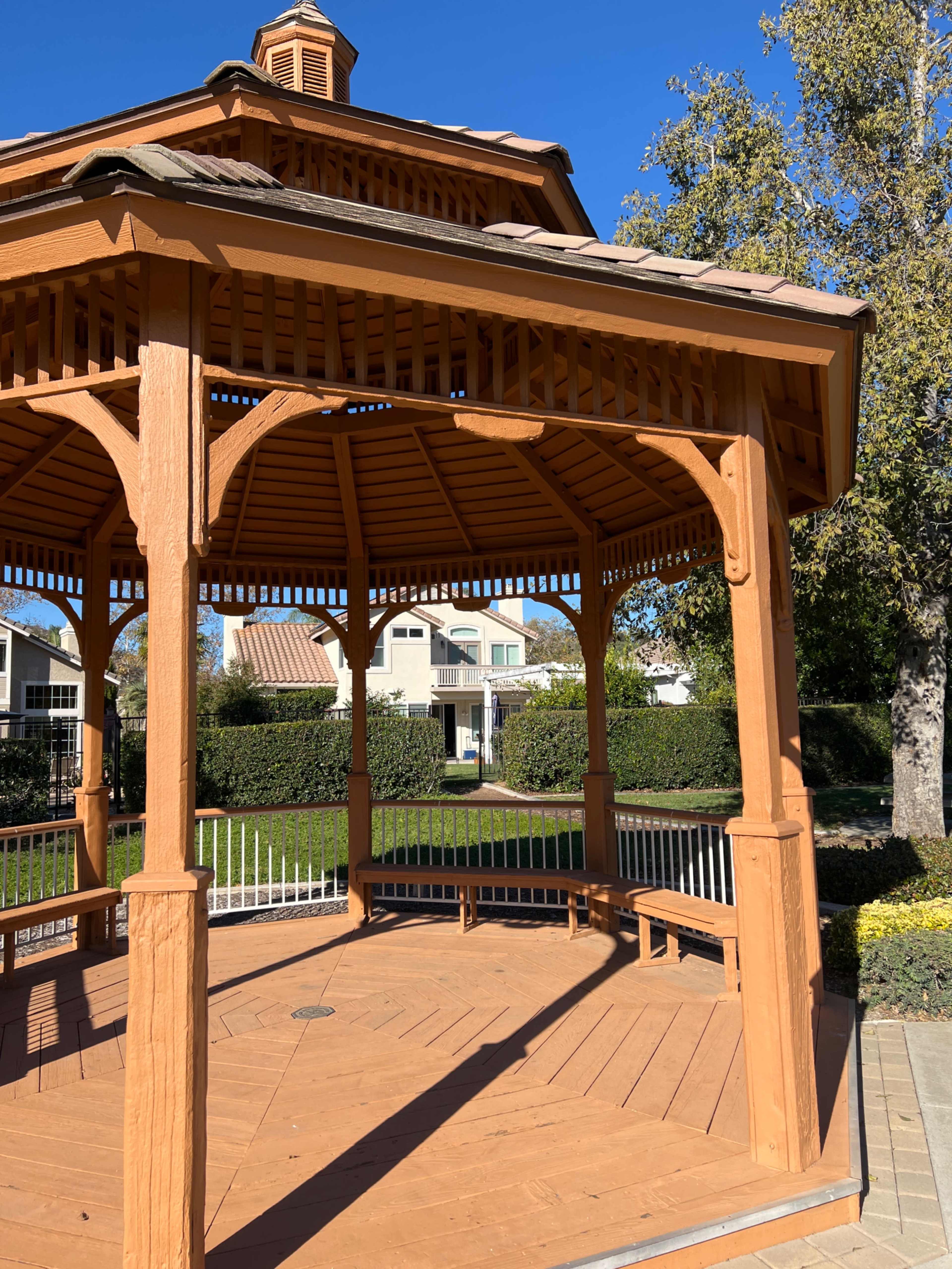 A wooden gazebo with a shingled roof stands in a park, surrounded by neatly trimmed hedges and nearby houses.