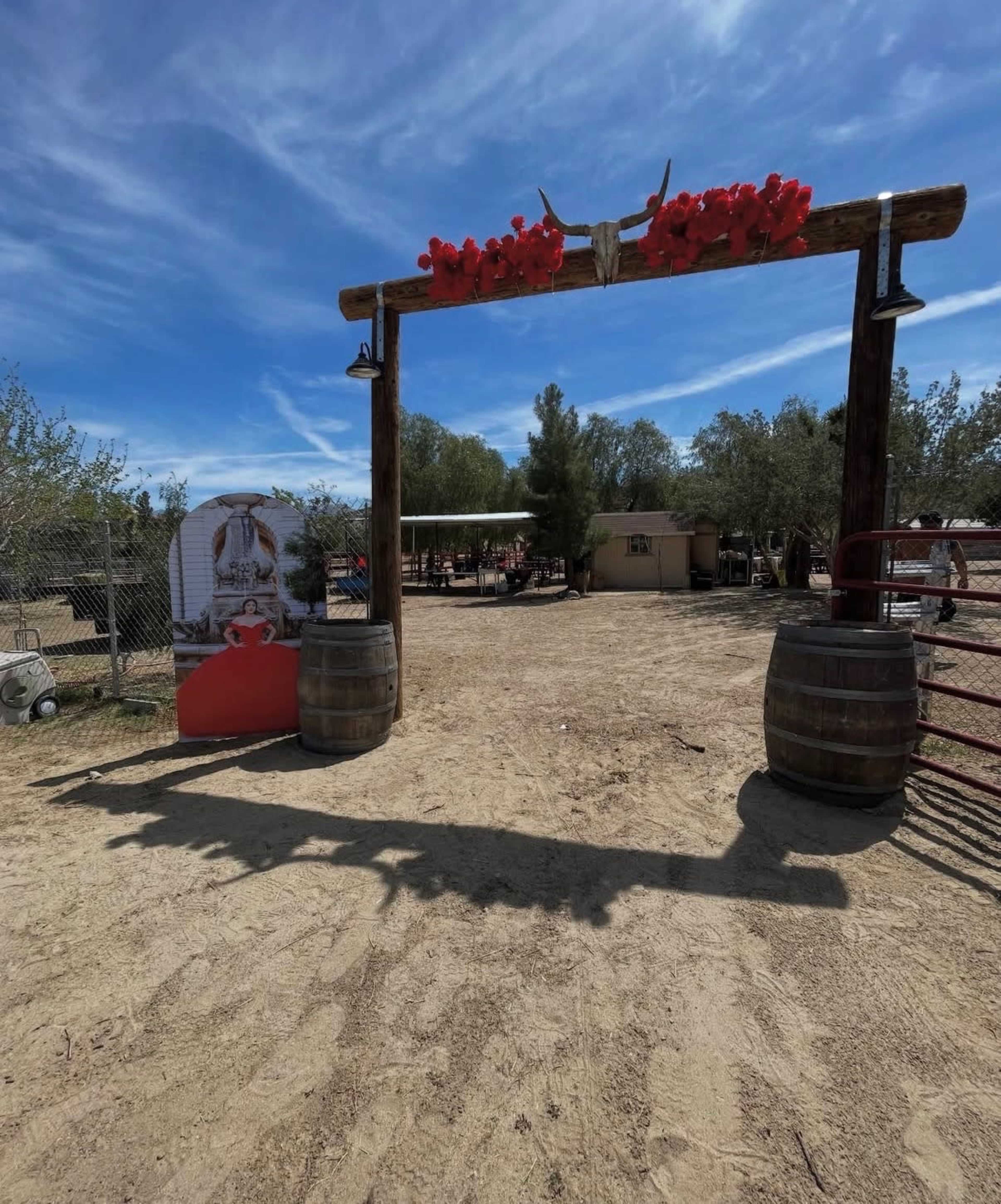 The image shows a wooden arch decorated with red flowers and a pair of horns, leading into a sandy area with several structures and trees in the background.