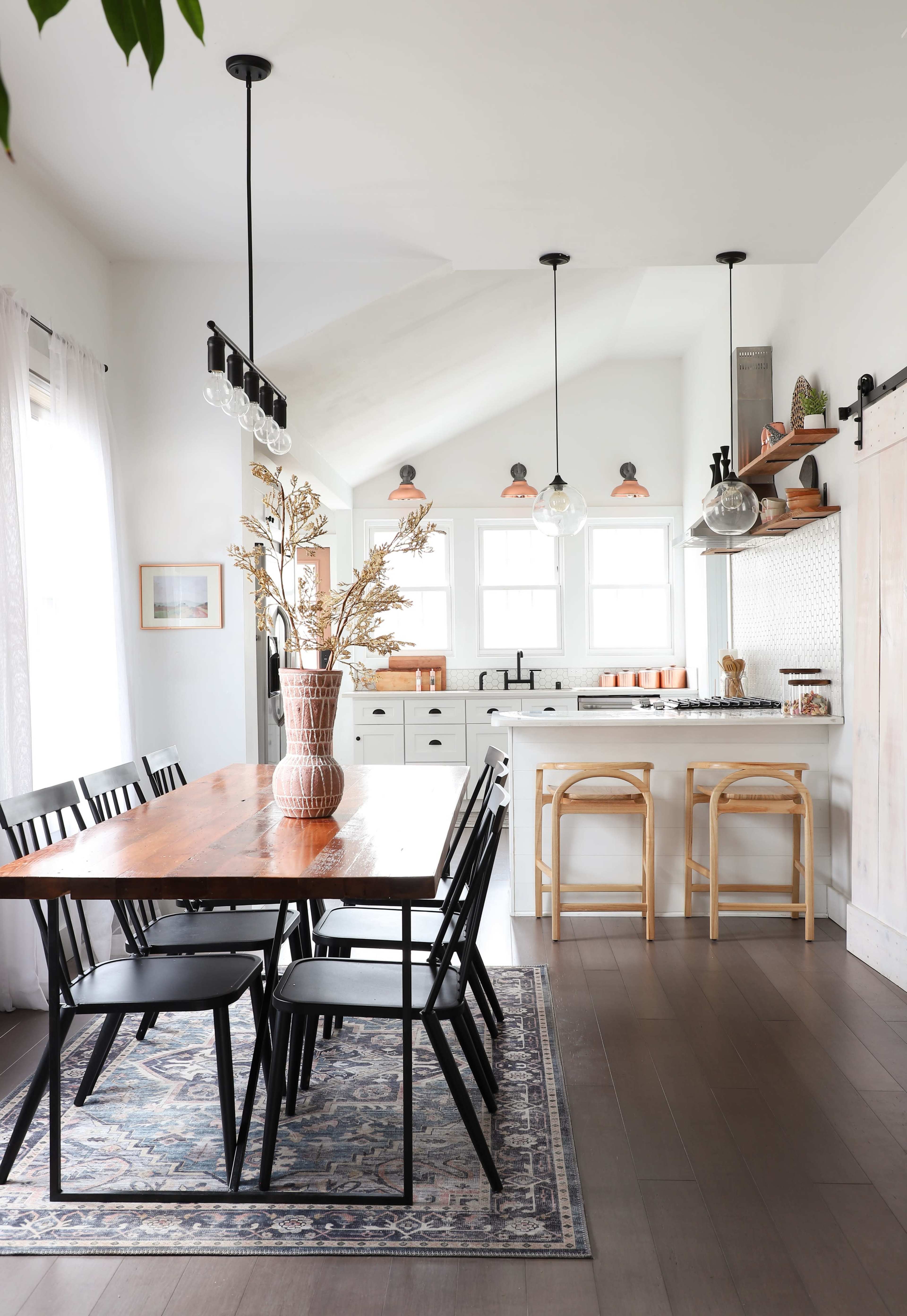 A modern kitchen features a wooden dining table surrounded by black chairs, with a bright decor including a vase of dried flowers and barstools at a counter.
