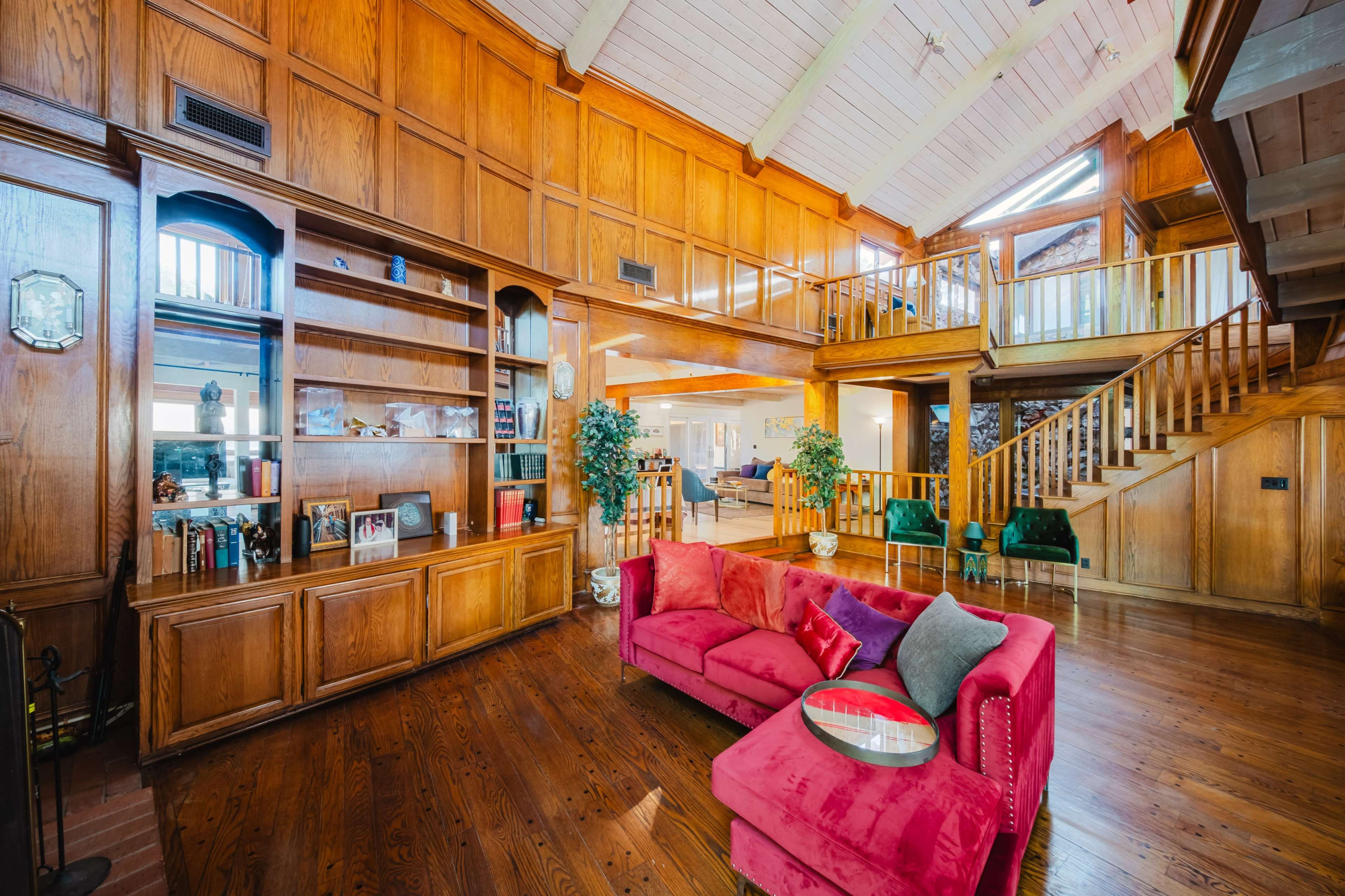 A spacious living room features wooden walls, a high ceiling with beams, a staircase, and a bright seating area with a pink sectional sofa.