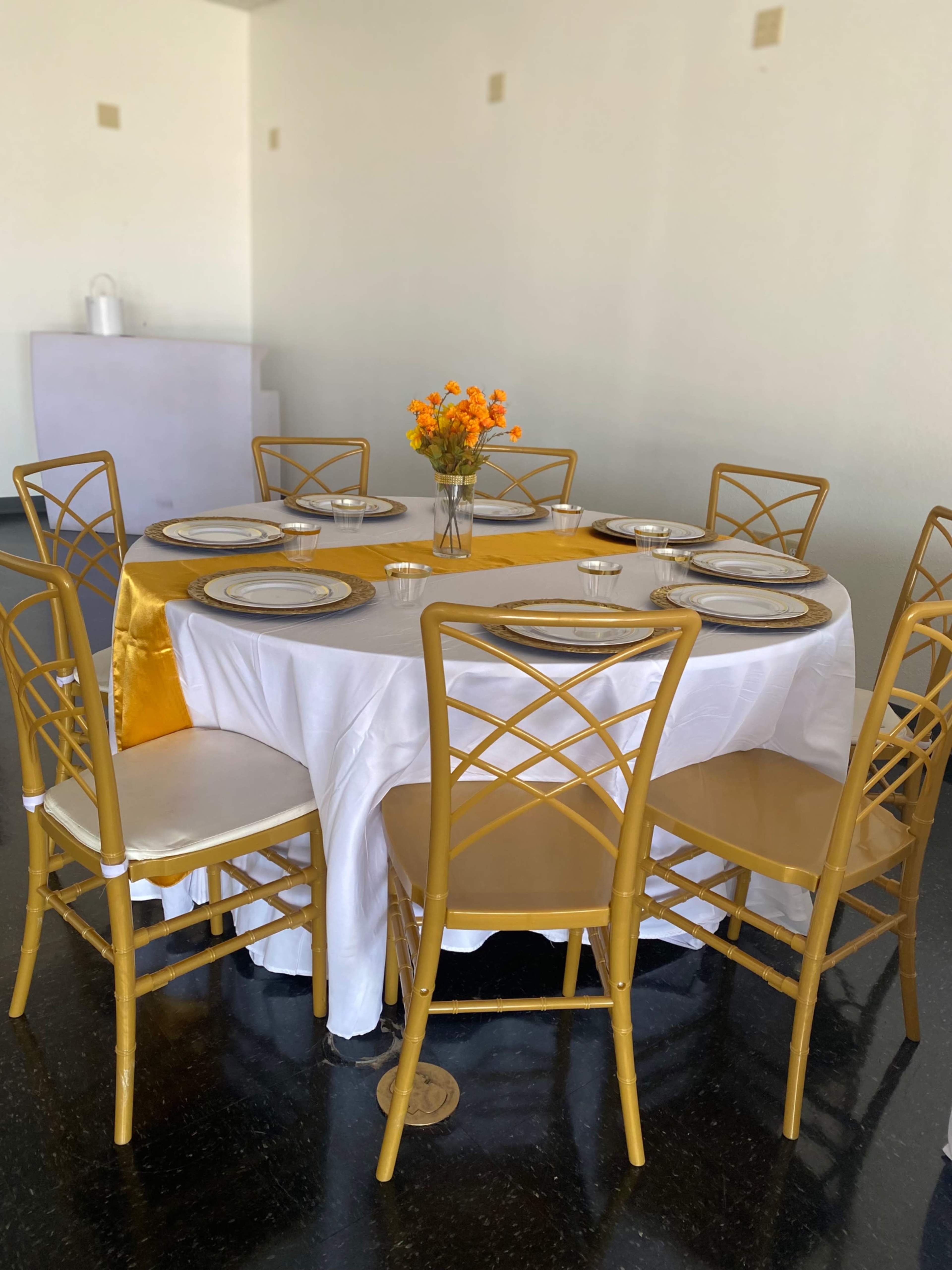 A round dining table with a white tablecloth and gold accents is set with plates and a vase of orange flowers in the center, surrounded by six gold chairs.