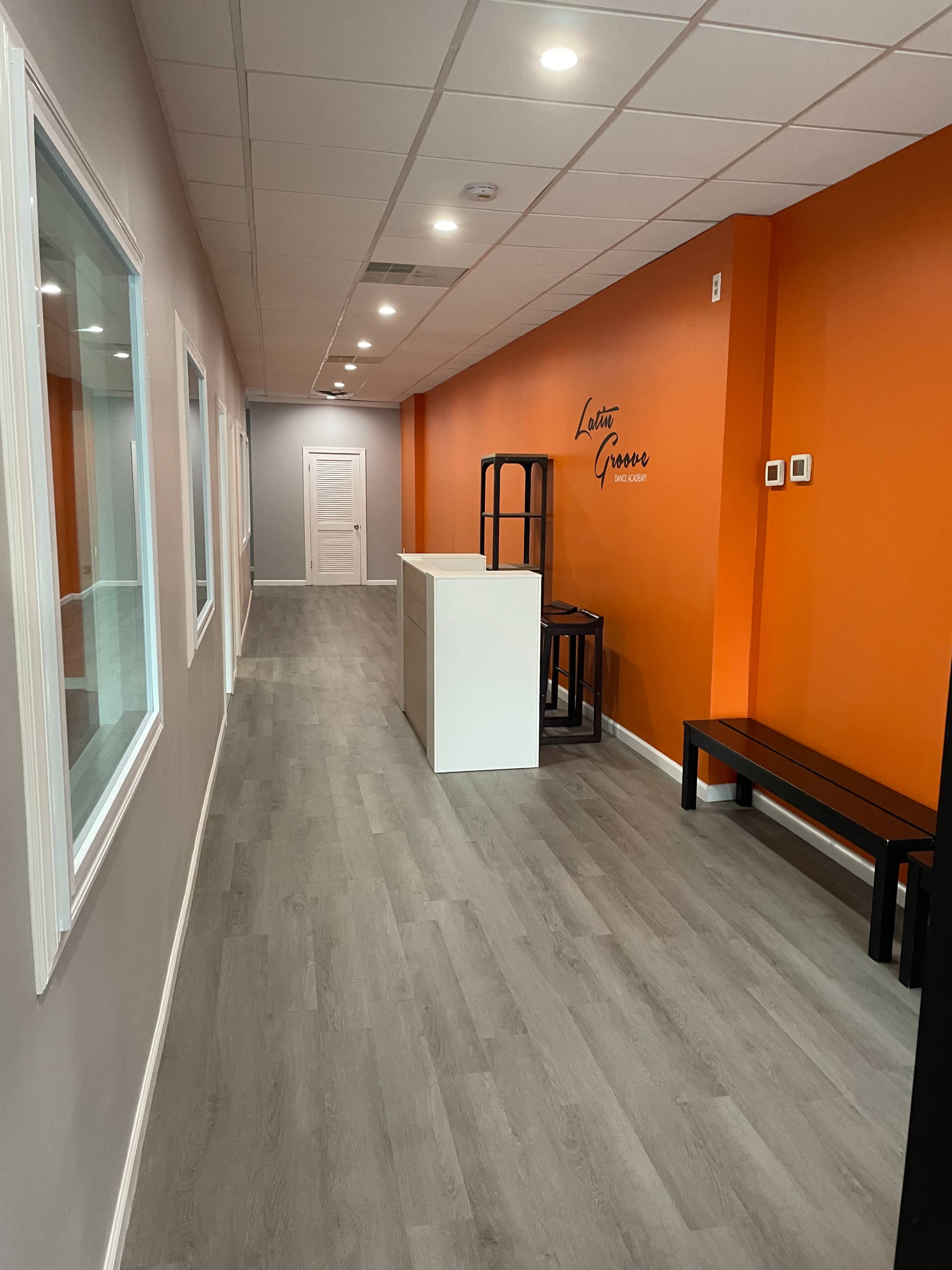 The image shows a narrow hallway with gray walls and an orange accent wall, featuring a reception area with a white counter and dark seating along one side.