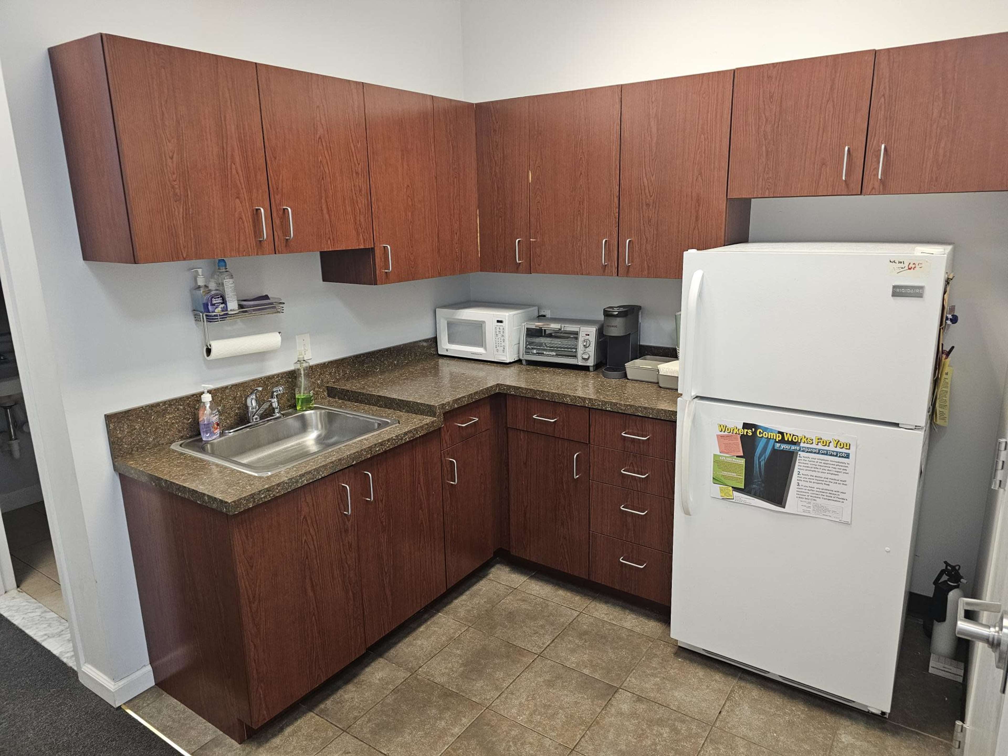 A kitchenette with wooden cabinets, a stainless steel sink, a microwave, a toaster oven, and a white refrigerator against a tiled floor.