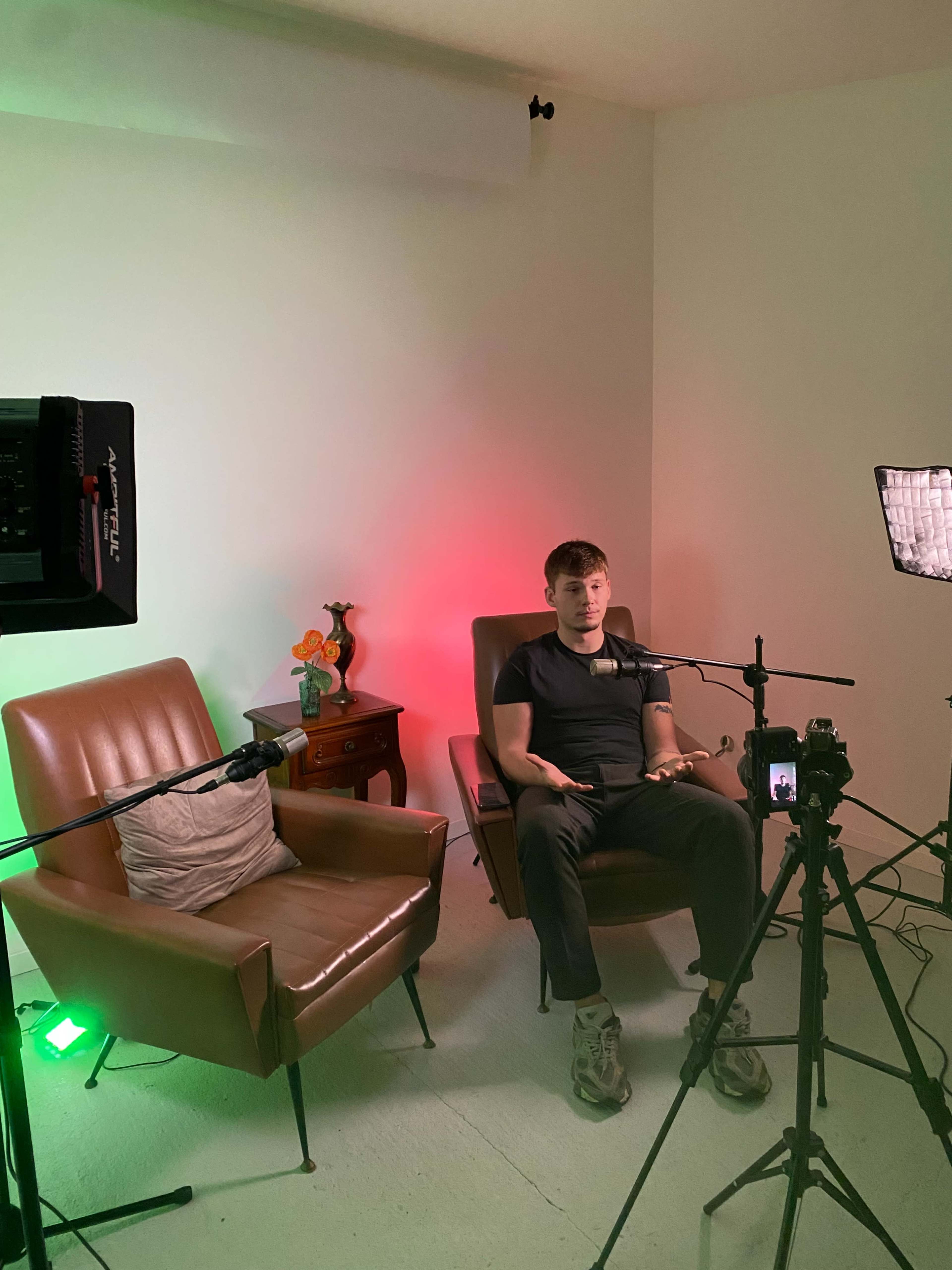 A young man sits in a chair in a well-lit room, flanked by two microphones, with two empty chairs and a small table in the background.