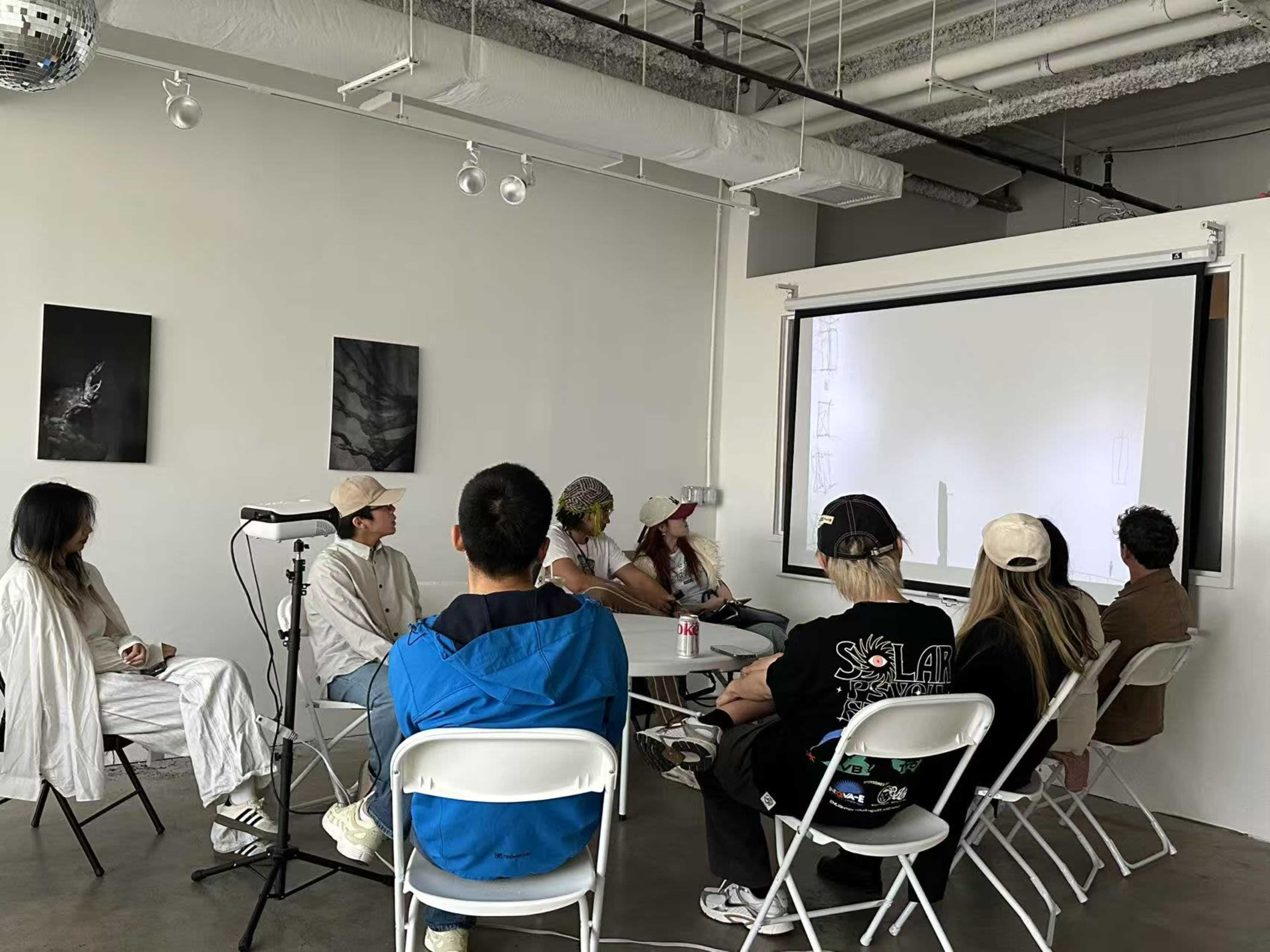 A group of people sits in chairs facing a large screen in a minimalist room with artworks on the walls.