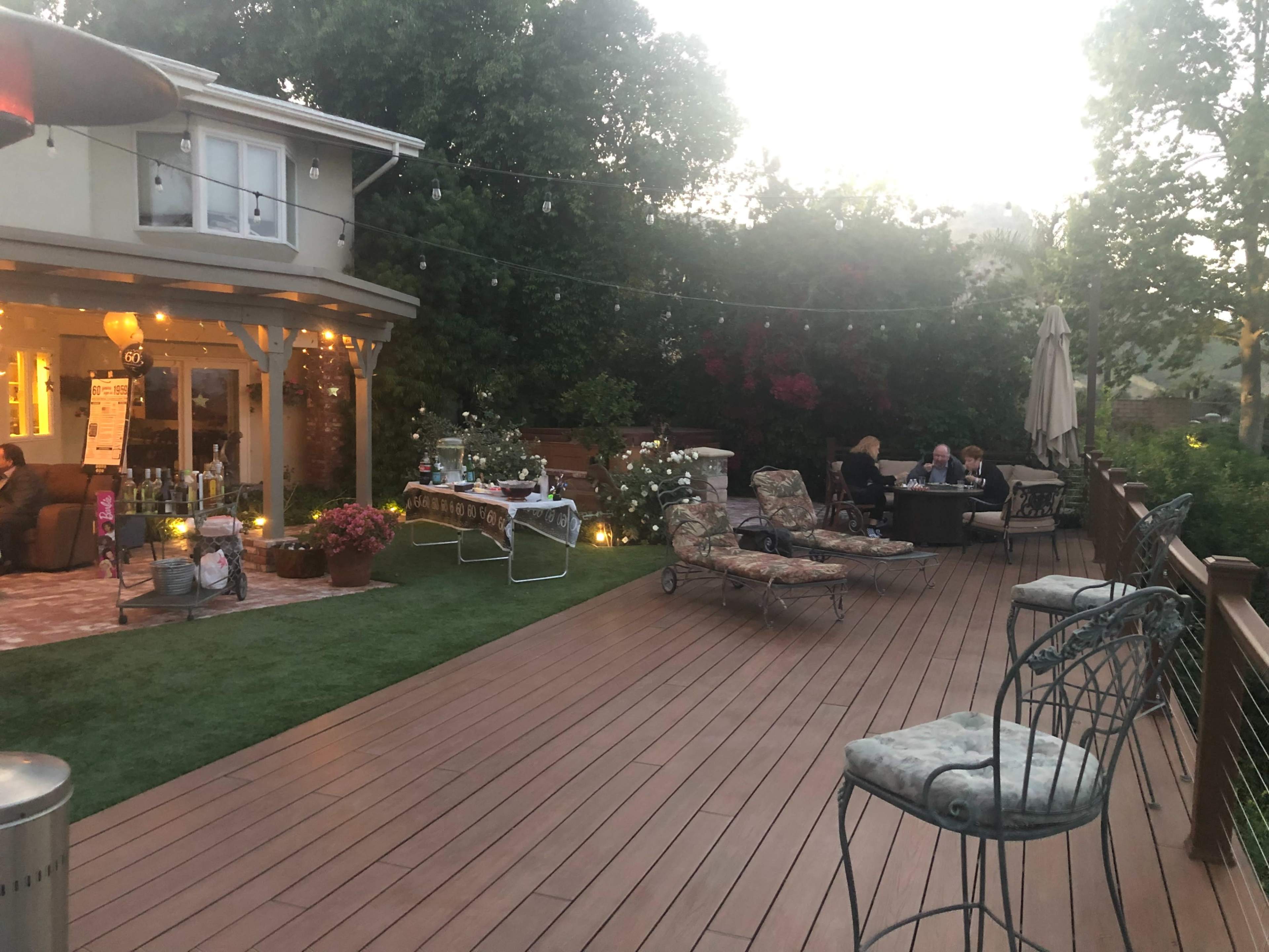A backyard patio with a wooden deck, outdoor seating, and string lights, featuring people socializing near a house surrounded by greenery.