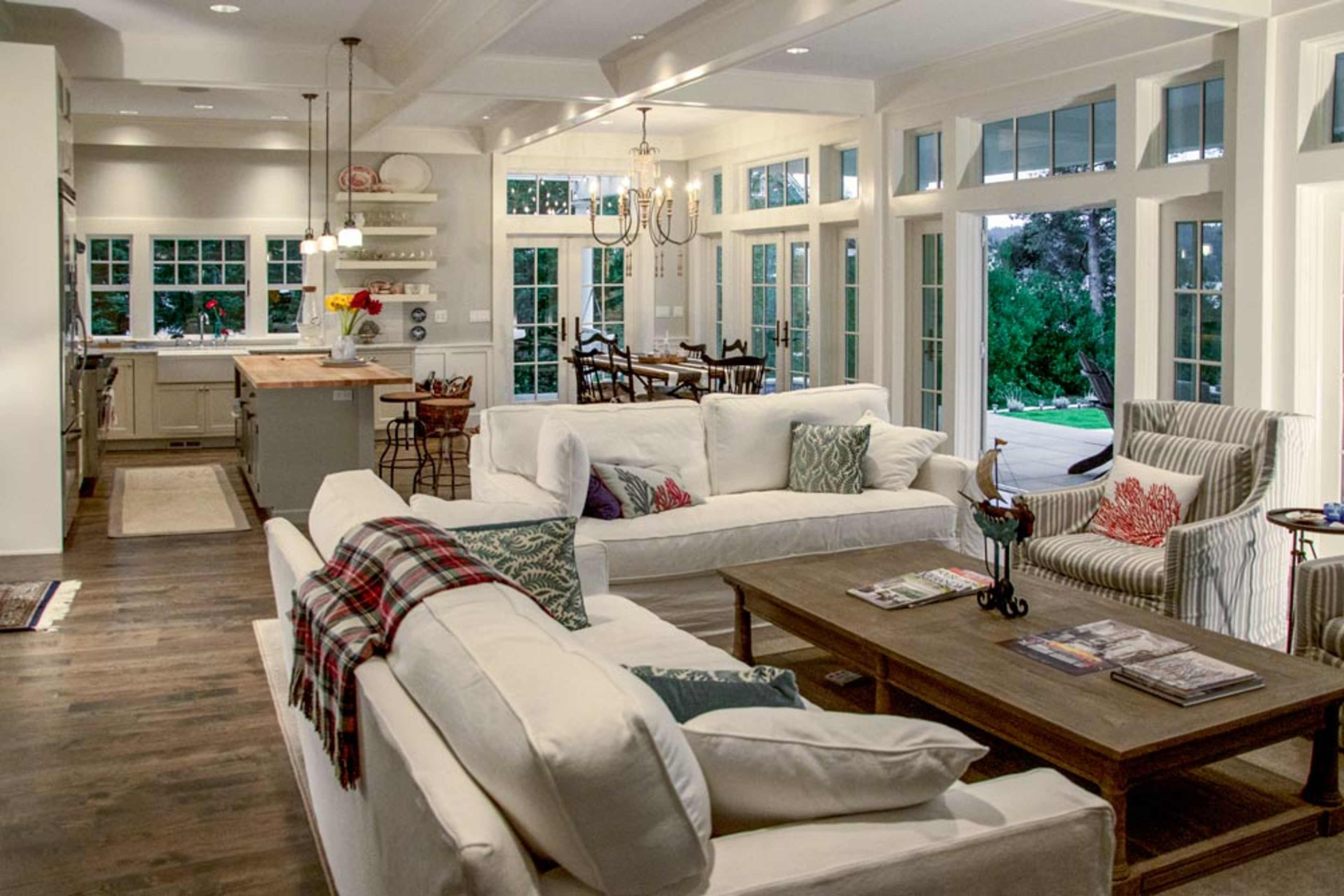 A spacious, well-lit living room with white sofas, a wooden coffee table, and large windows that overlook an outdoor area.
